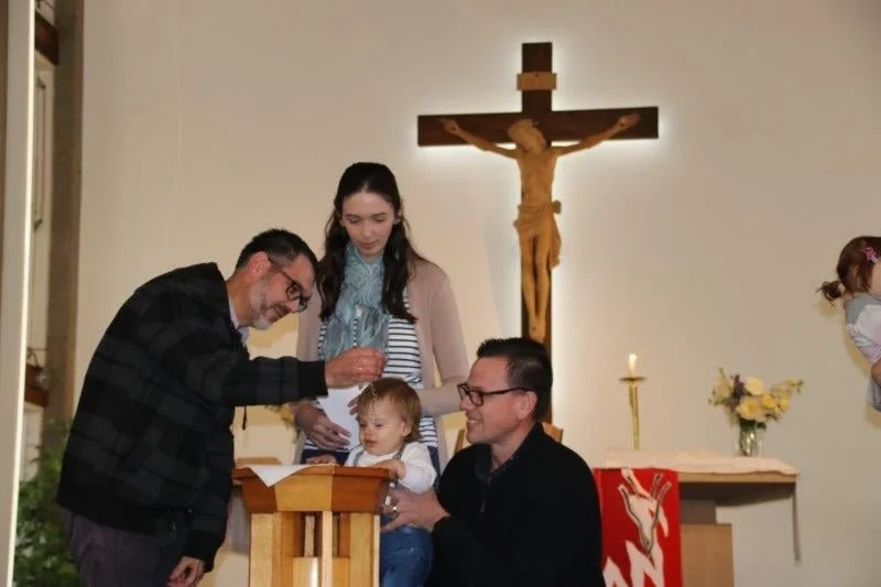 Family gathered at a baptism or religious ceremony inside a church, with a cross on the wall behind them, a priest or officiant, and a young child in front of a baptismal font.