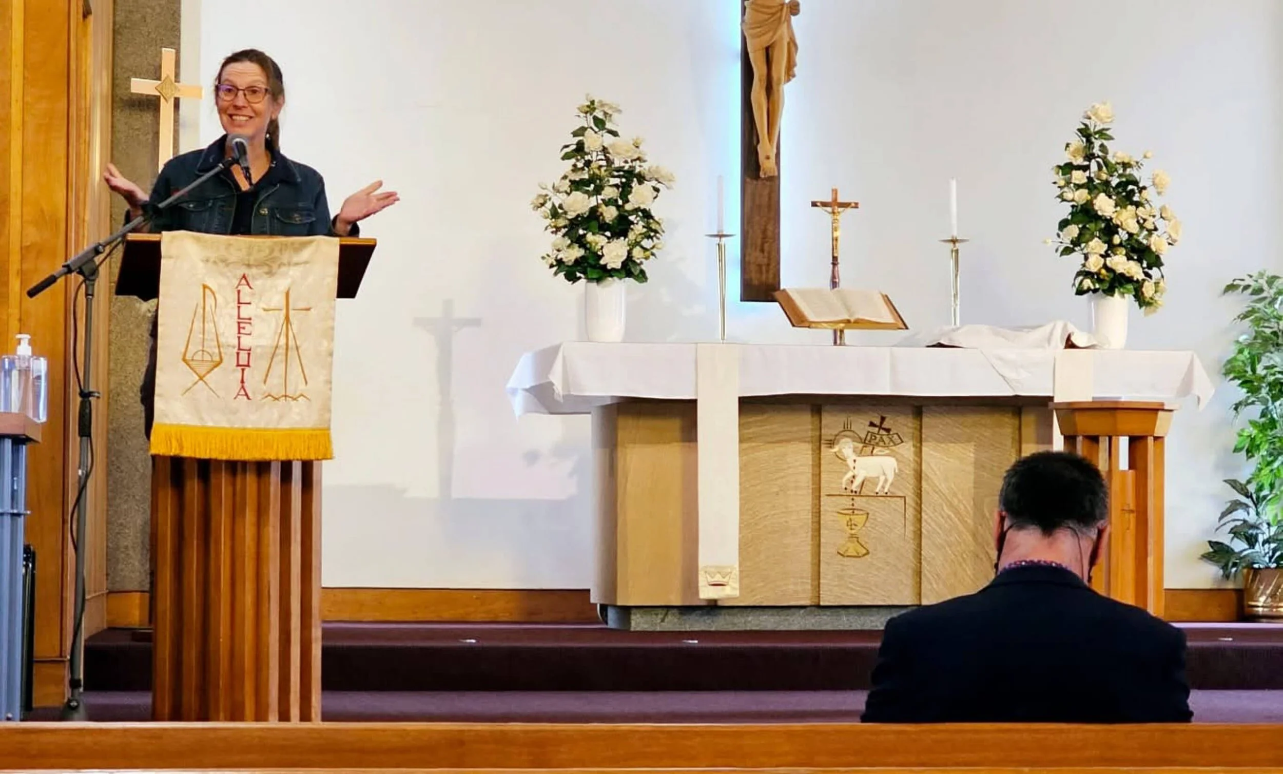 A woman standing at a lectern with a microphone, smiling, inside a church. The church altar has flowers, candles, a crucifix, an open book, and religious icons, including a hanging crucifix and a wooden panel with Christian symbols.