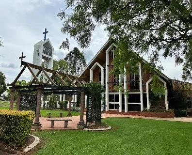 A church with a sloped roof and large windows, surrounded by trees and a well-maintained lawn, with a stone cross on top of a small white bell tower.
