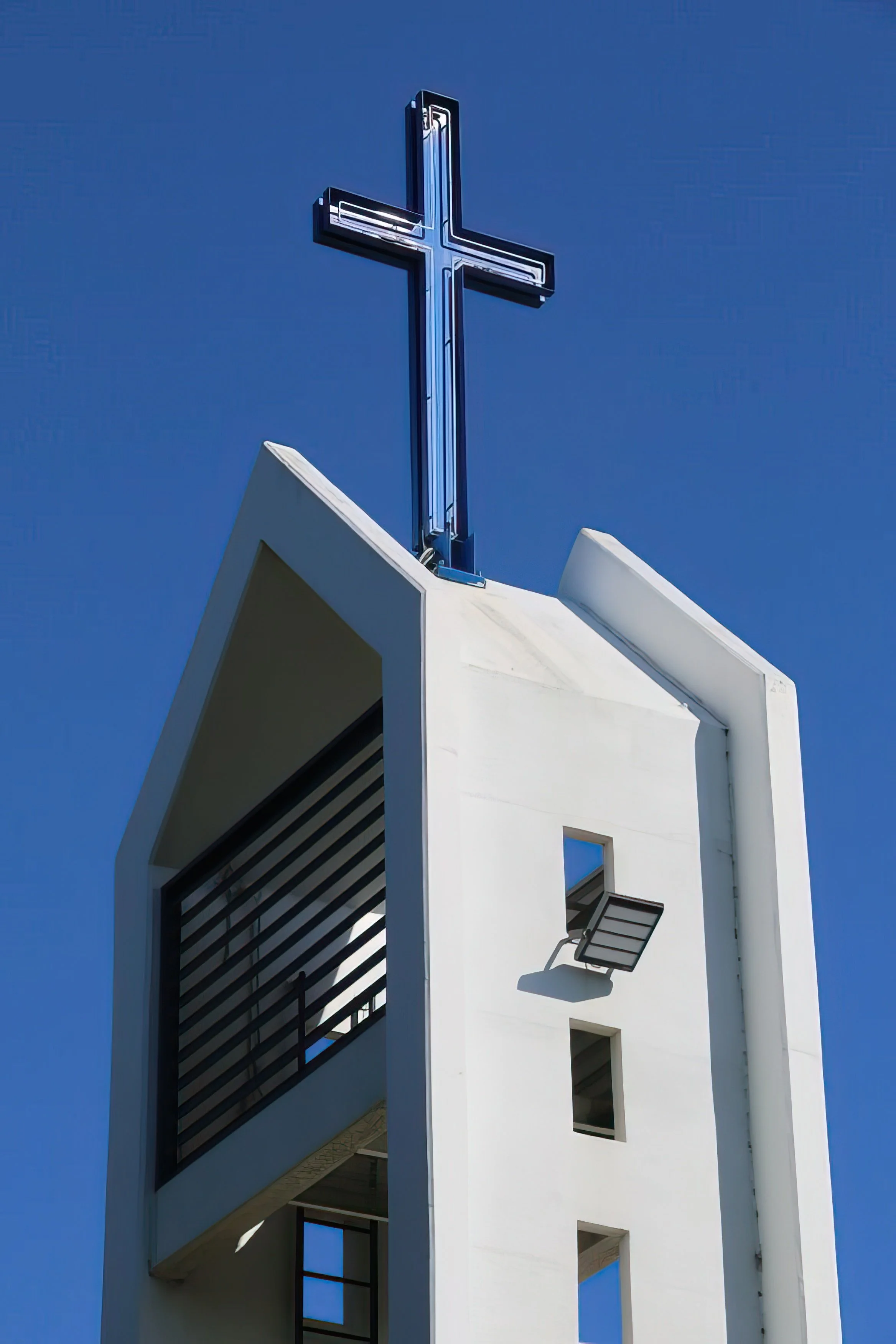 A modern white building with a cross-shaped blue neon-lit sign on top, a large overhanging balcony with black horizontal slats, and several windows against a clear blue sky.