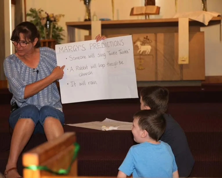 A woman sits on a step holding a large piece of paper titled "Marcy's Predictions" with three bullet points about someone singing, a rabbit, and rain, as two young boys sit on the floor looking at her in a church or community setting.