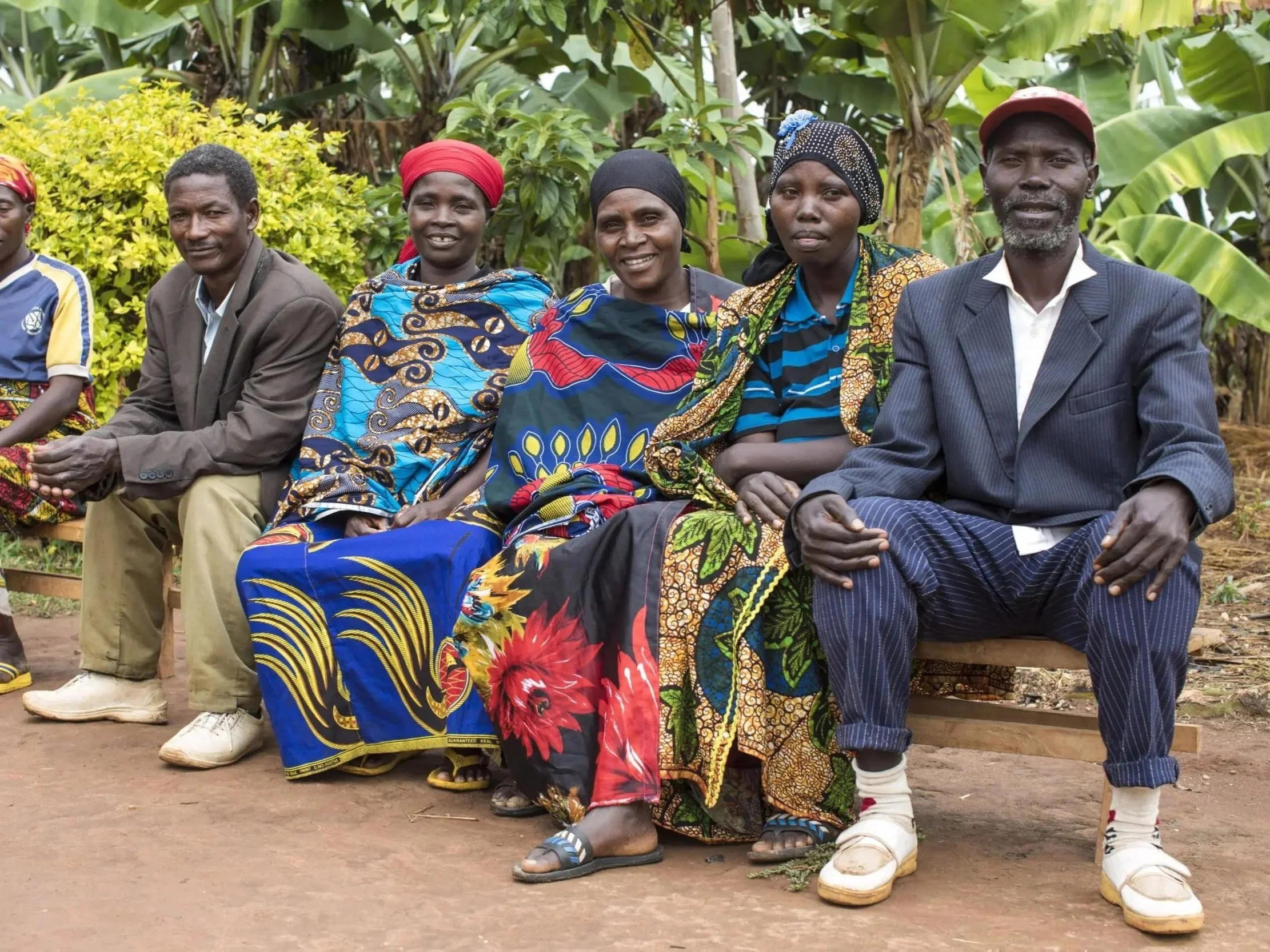 Group of African people sitting outdoors in front of lush green bushes and trees, dressed in colorful traditional and modern clothing.