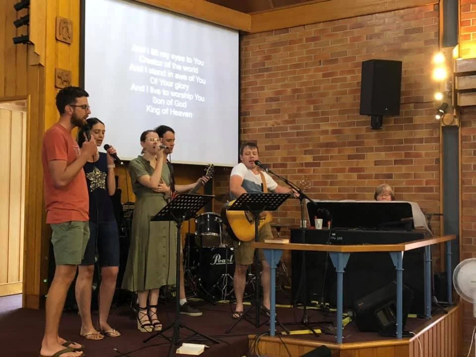 A group of six people singing and playing music on a stage with a brick wall background, a projector screen displaying song lyrics, and musical instruments including drums, a keyboard, and a guitar.