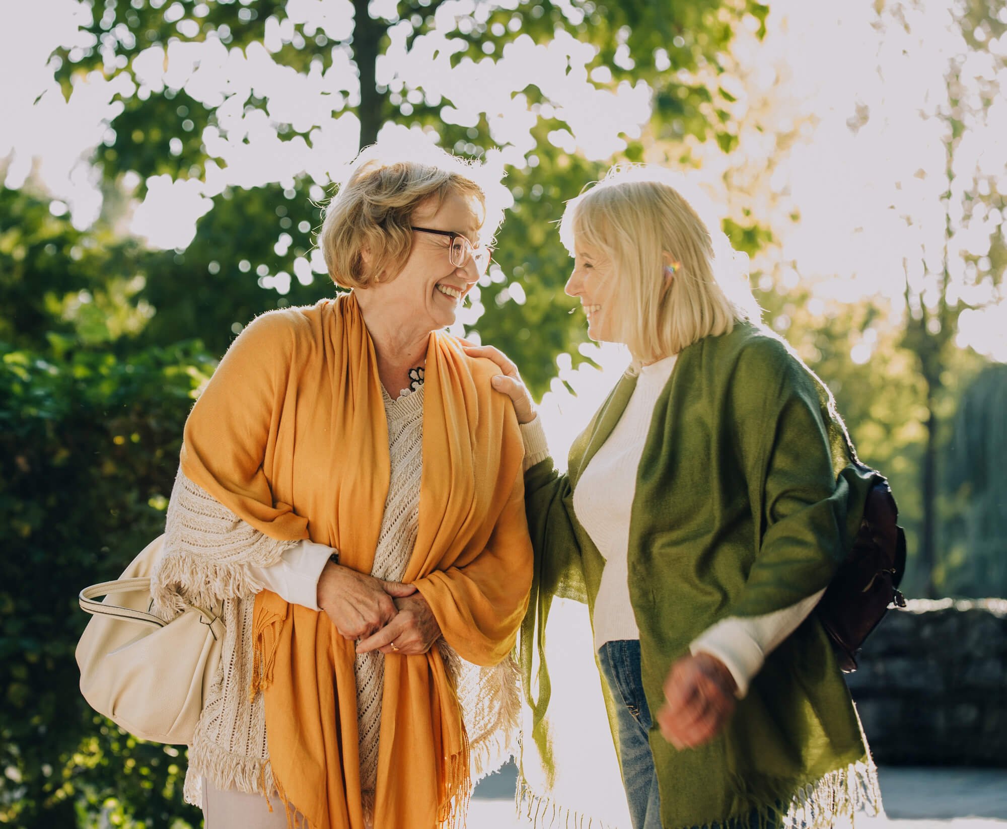 Two women smiling and interacting outdoors in a sunny park, with trees and sunlight in the background.
