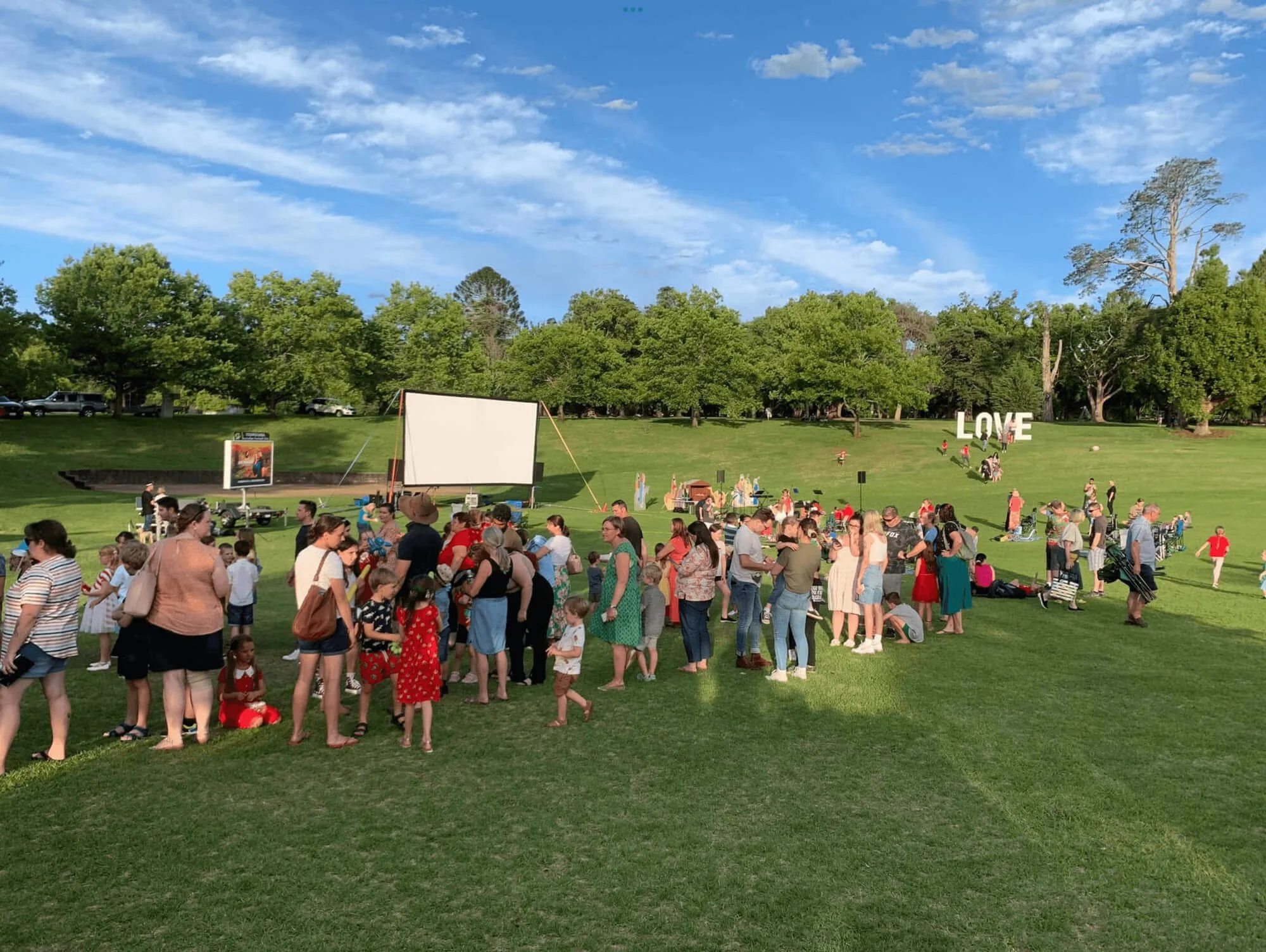 A large outdoor gathering in a park with many people, some standing and others sitting on the grass, under a blue sky with scattered clouds. There are trees in the background and a large 