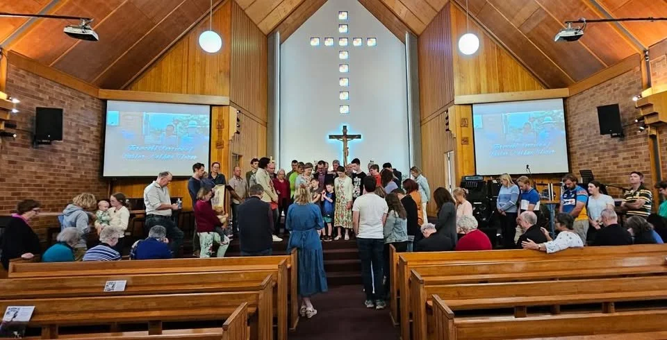 People gather in the front of a church altar for a religious ceremony. The church has wooden walls, a large cross on the wall, and two large screens displaying images. Several people are seated in pews, and some are standing or kneeling near the alta