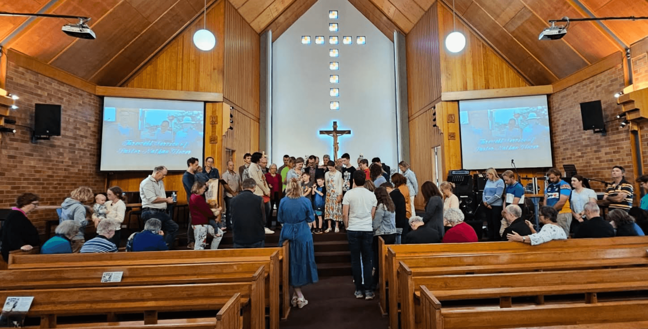 A church congregation gathered in a wooden church interior with a crucifix on the wall, during a service or ceremony, with people standing in the front, some holding or reading from books, and a large congregation seated in pews.