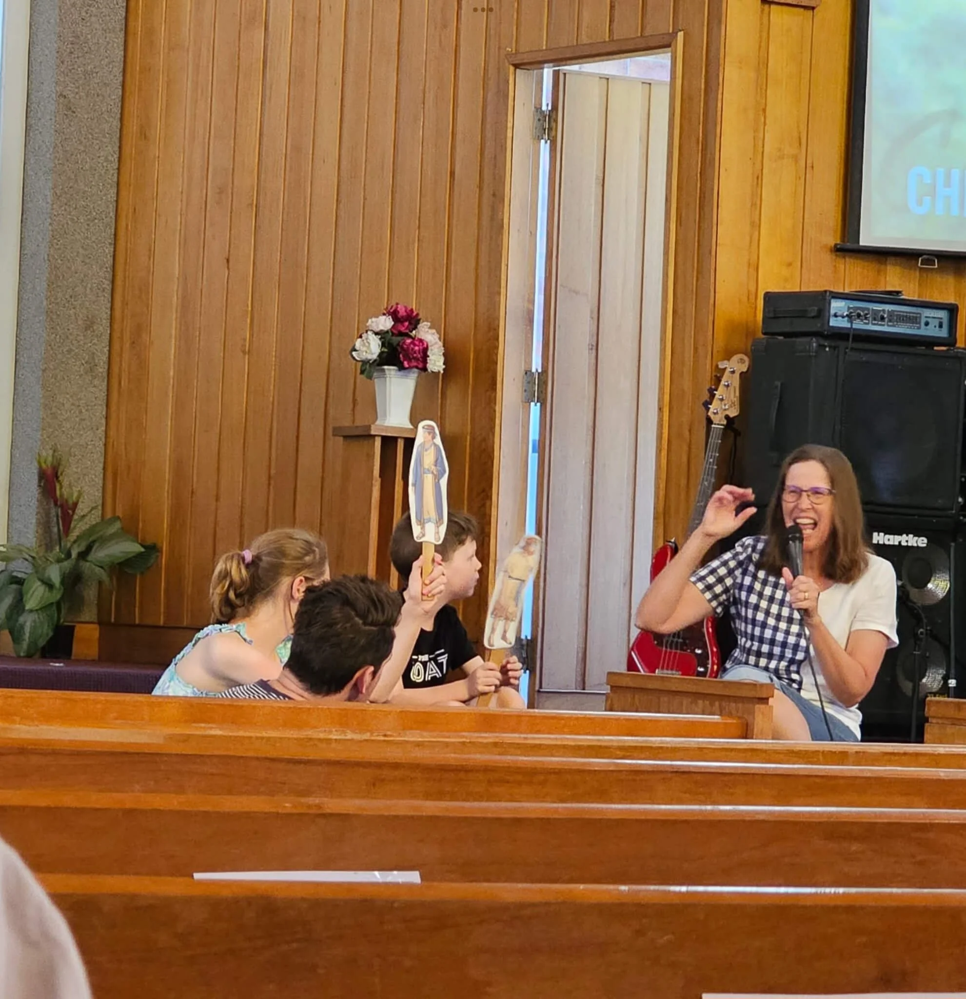 A woman in a checkered shirt and glasses holding a microphone and smiling. Several children sitting at a table, some holding paper cutouts of religious figures. The background has a wooden wall with a vase of flowers and a religious picture with a bl