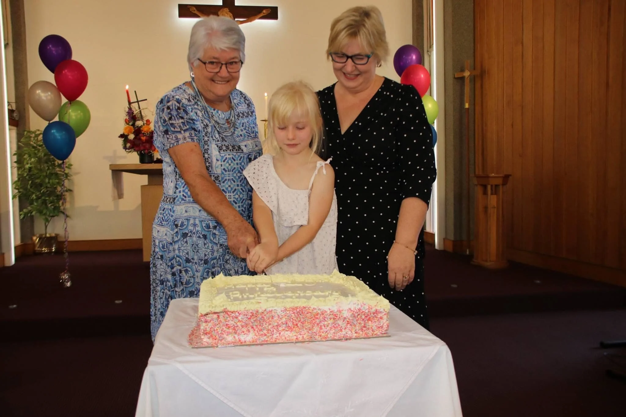A young girl with blonde hair, wearing a white dress with pink accents, stands at a table with a large decorated cake. Two older women with glasses stand on either side of her, smiling and looking down at her. The scene is in a room decorated with ba