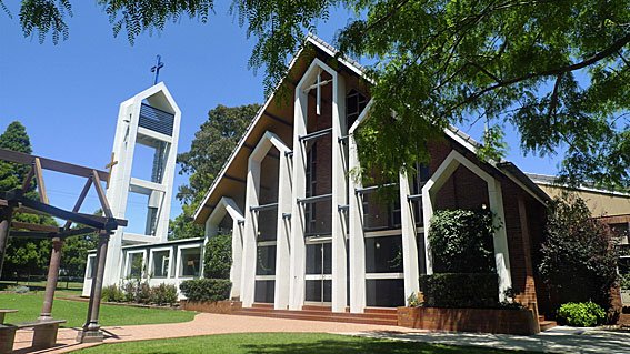 Modern church building with tall, angled windows, a white steeple with a cross at the top, surrounded by greenery and trees.