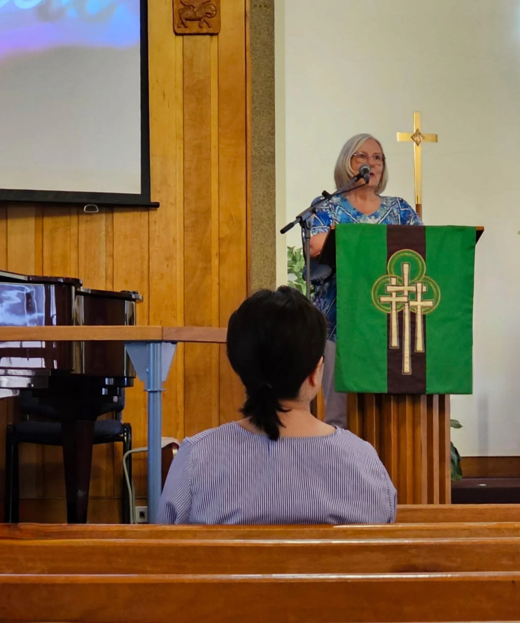 A woman with gray hair and glasses speaking at a church podium. She is wearing a blue patterned top and behind her is a gold cross on a white wall with wooden paneling.