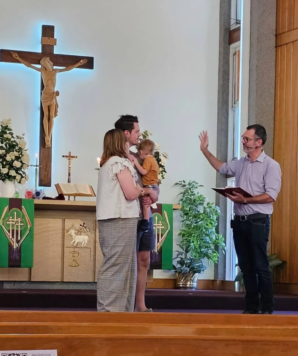 A family participating in a baptism or christening service inside a church, with a priest or minister officiating, at the altar with a crucifix and religious symbols in the background.