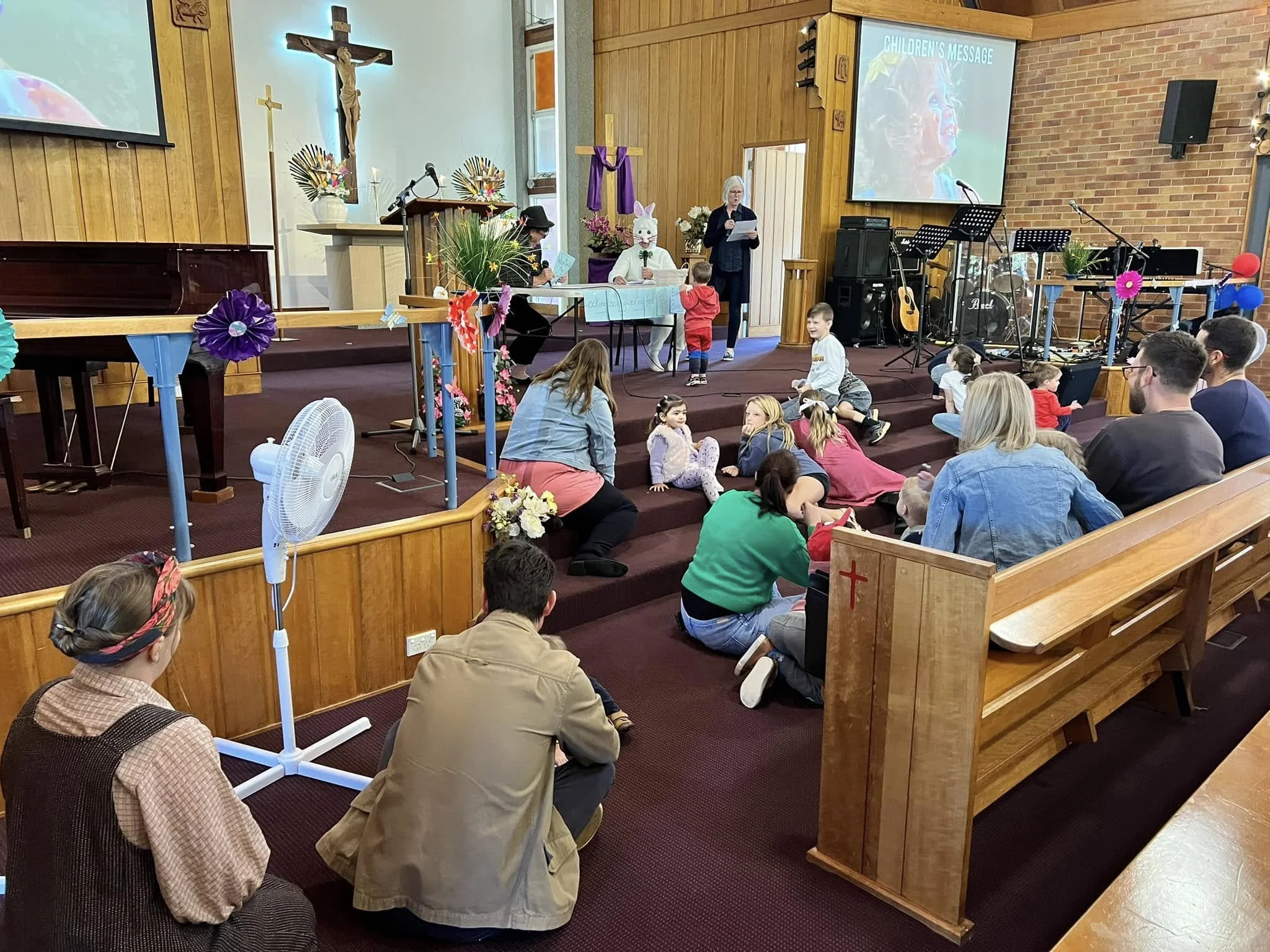 Children's message at a church service with children sitting on the floor and a woman reading papers near bunny and lamb costumes, with religious icons and a cross in the background, and a large screen displaying 'Children's Message'.