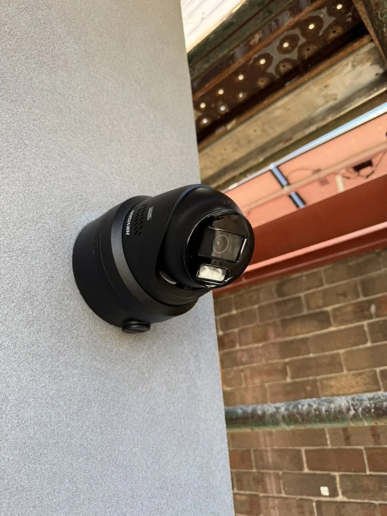 A black Hikvision security camera with a motorized zoom lens, mounted on a gray surface outdoors, with a brick wall and a metal pipe in the background.