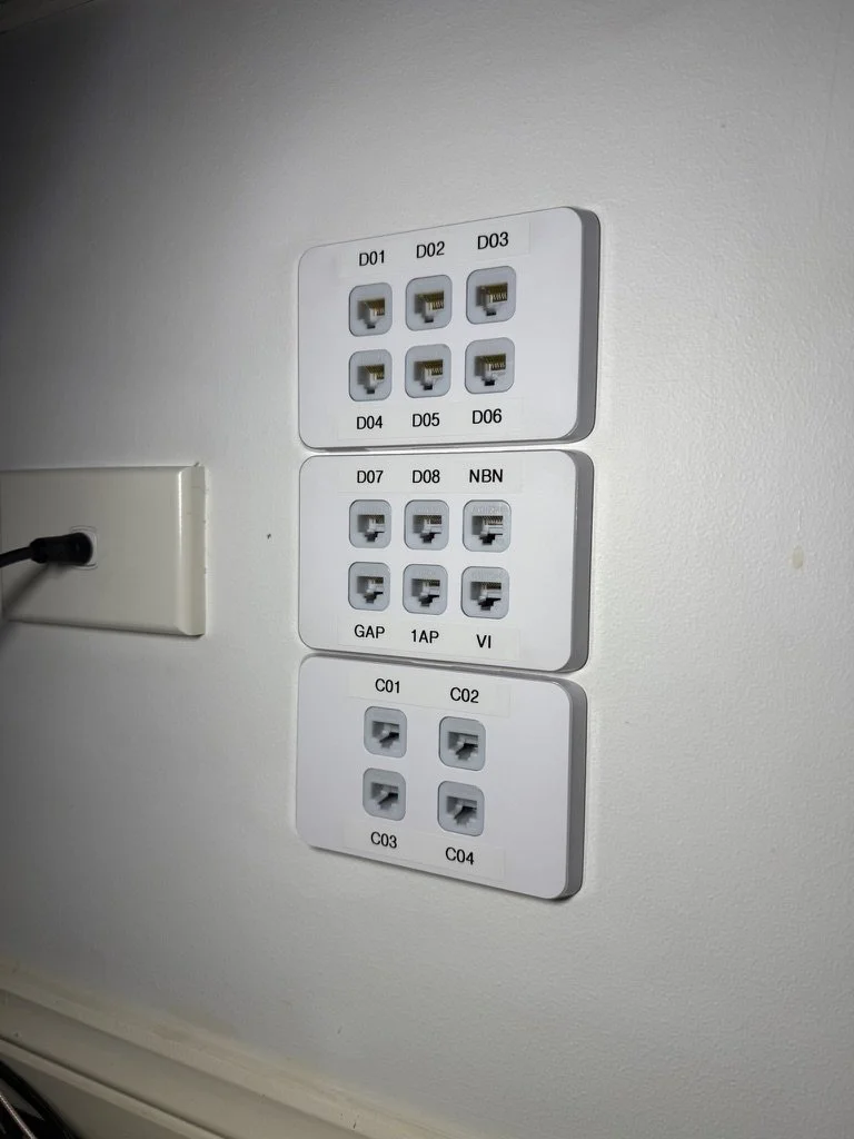 Wall-mounted network socket panel with multiple ports labeled D01 to D08, NBN, GAP, 1AP, VI, and C01 to C04, next to a TV outlet with a black plug.