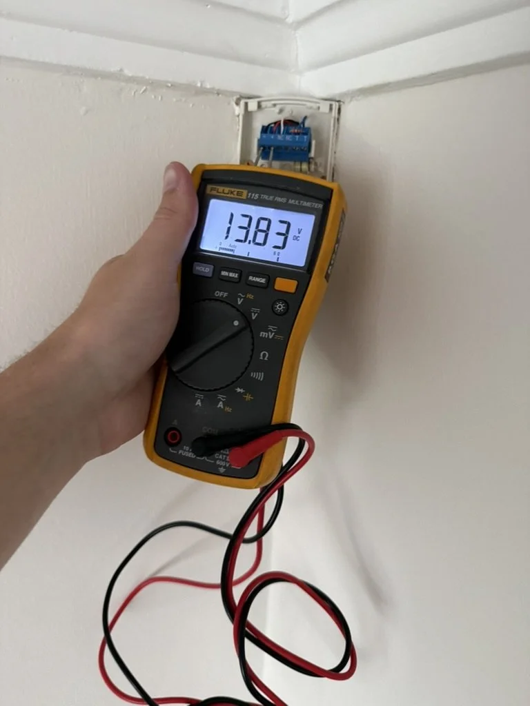 A person holding a digital multimeter measuring 13.83 volts DC on a Bosch PIR.