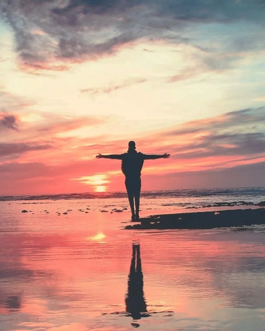 A person standing on a beach with arms outstretched during sunset, with their reflection visible in the water.