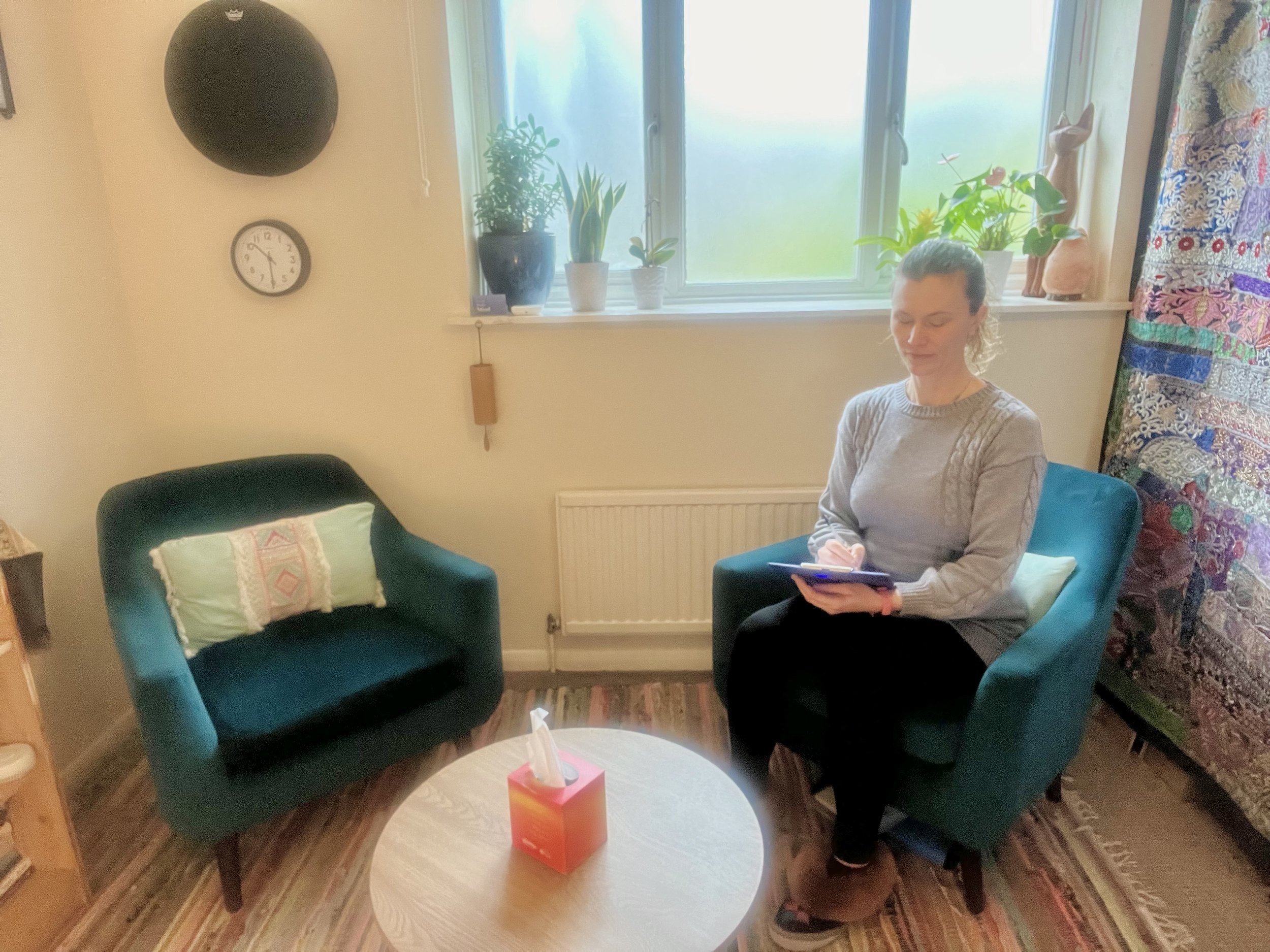 Ella sitting in her therapy room with a green armchair, small round table, and decorations. She is holding a clipboard and writing. There are plants on the windowsill, a wall clock, and colorful textiles in the setting.