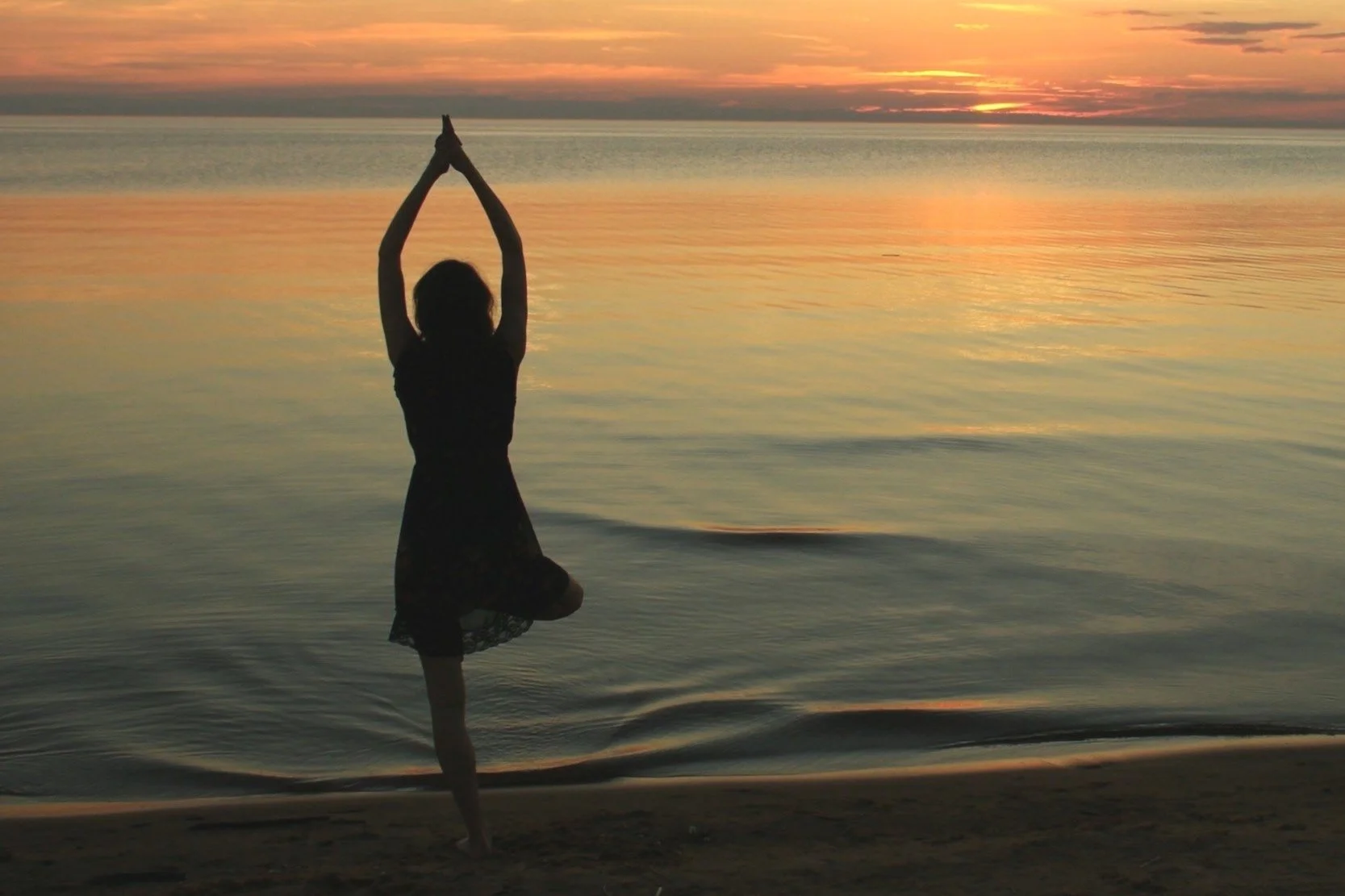 A woman practicing yoga on a beach during sunset, balancing on one leg with hands raised above her head in a prayer position.