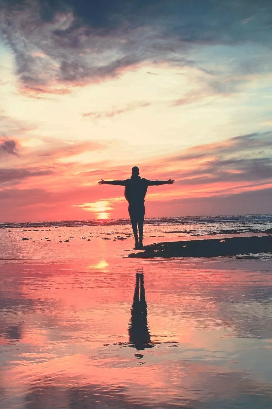 Silhouette of a person with arms outstretched at sunset, reflecting on wet sand by the ocean.