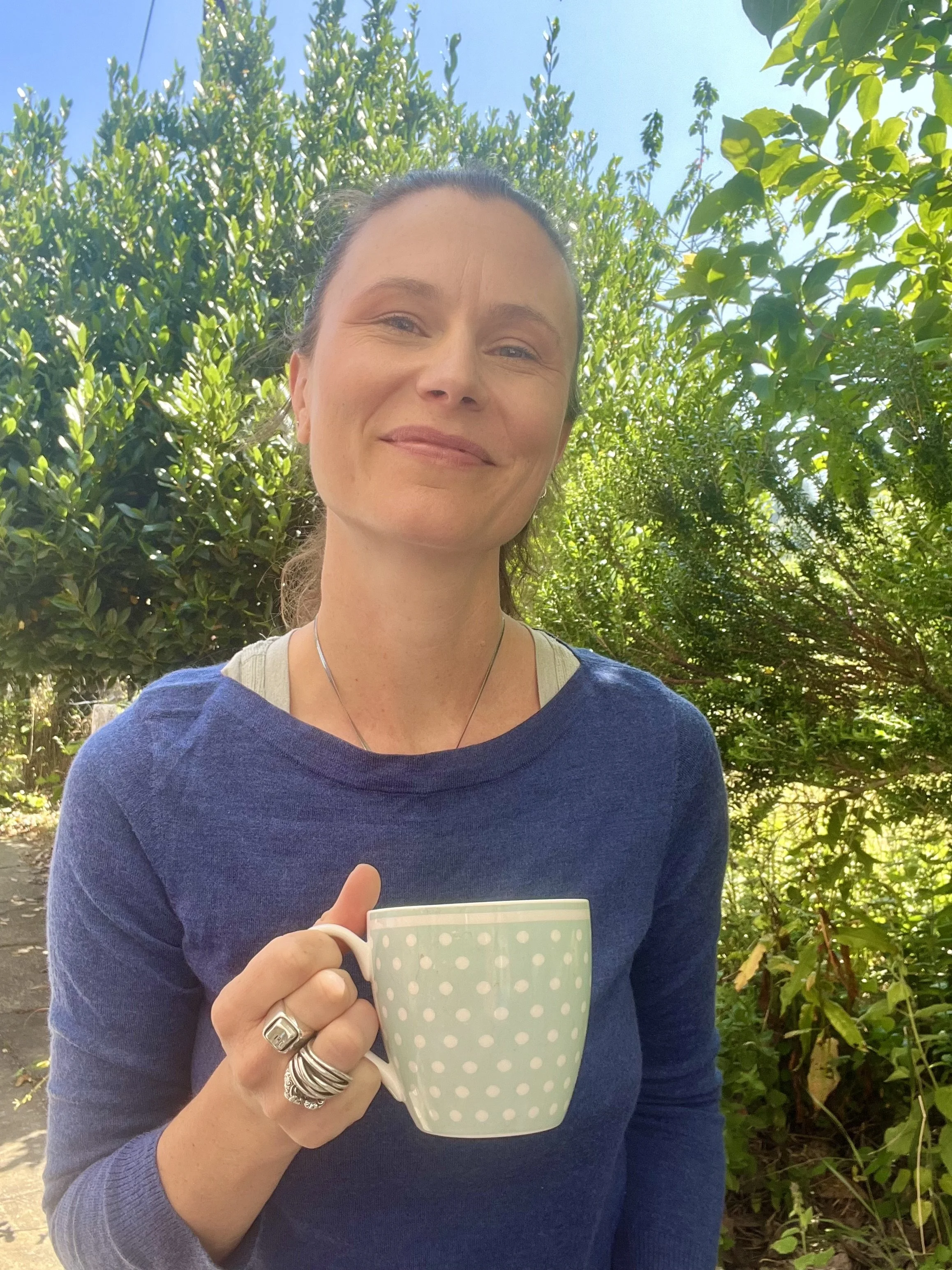 Ella smiling holding a green mug with trees in the background and a blue sky