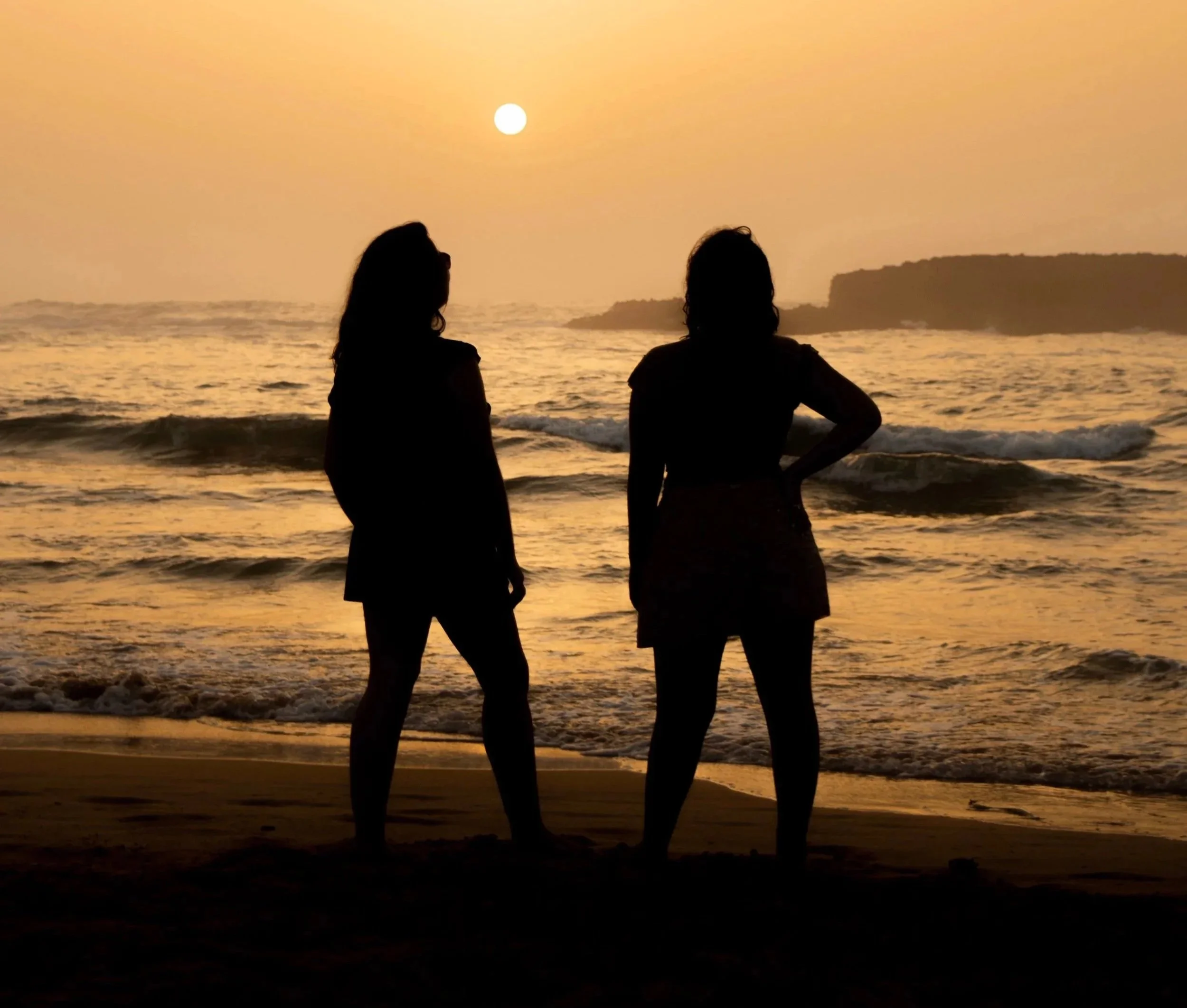 Two women standing on a beach during sunset, silhouetted against the orange sky and ocean, with a rocky landmass in the background.