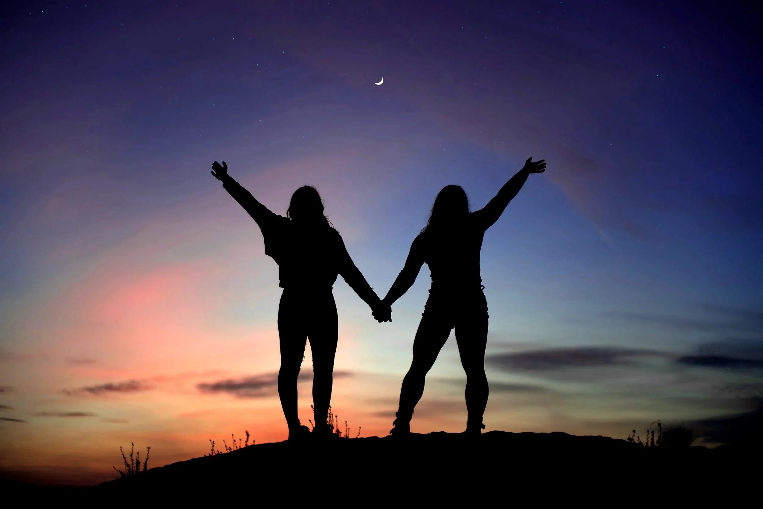Silhouette of 2 women holding hands with thier arms in the air and a colourful sky in the background
