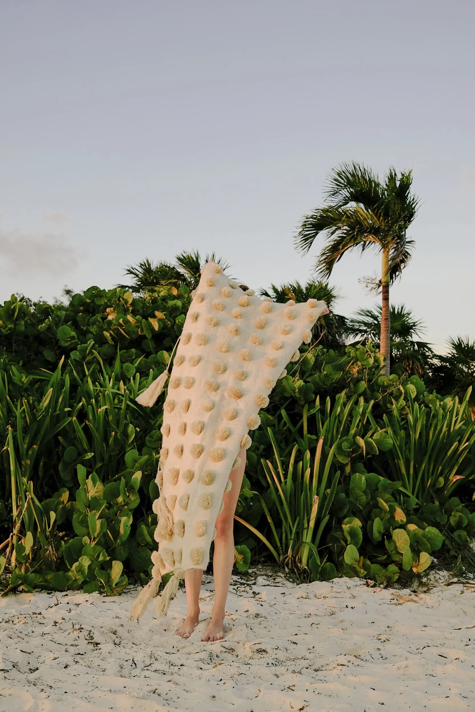 Personne portant une tunique en laine blanche texturée avec des pompons, debout sur une plage de sable avec des plantes tropicales en arrière-plan.