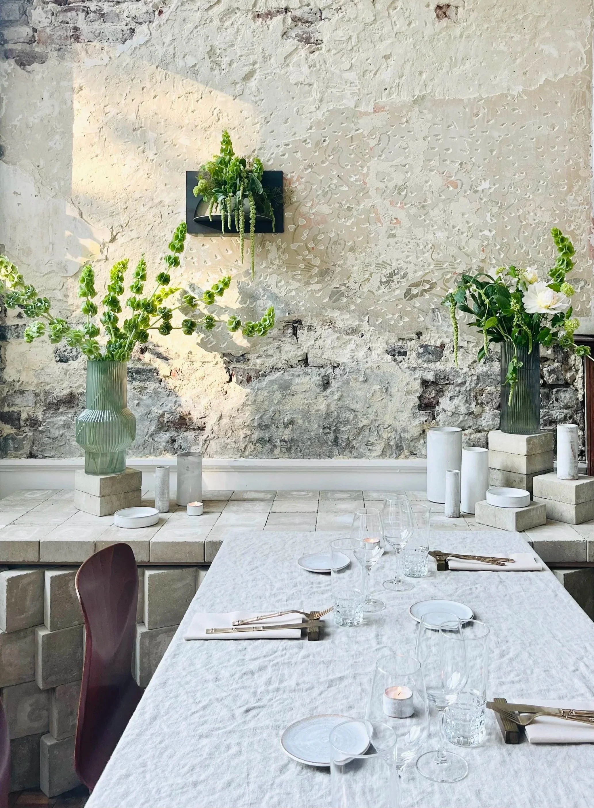 Une table élégante dressée dans une salle avec un mur en pierre. La table est couverte d'une nappe blanche, avec des verres à vin, assiettes, couverts, et bougies. Deux grands vases avec des fleurs vertes et blanches décorent la pièce.