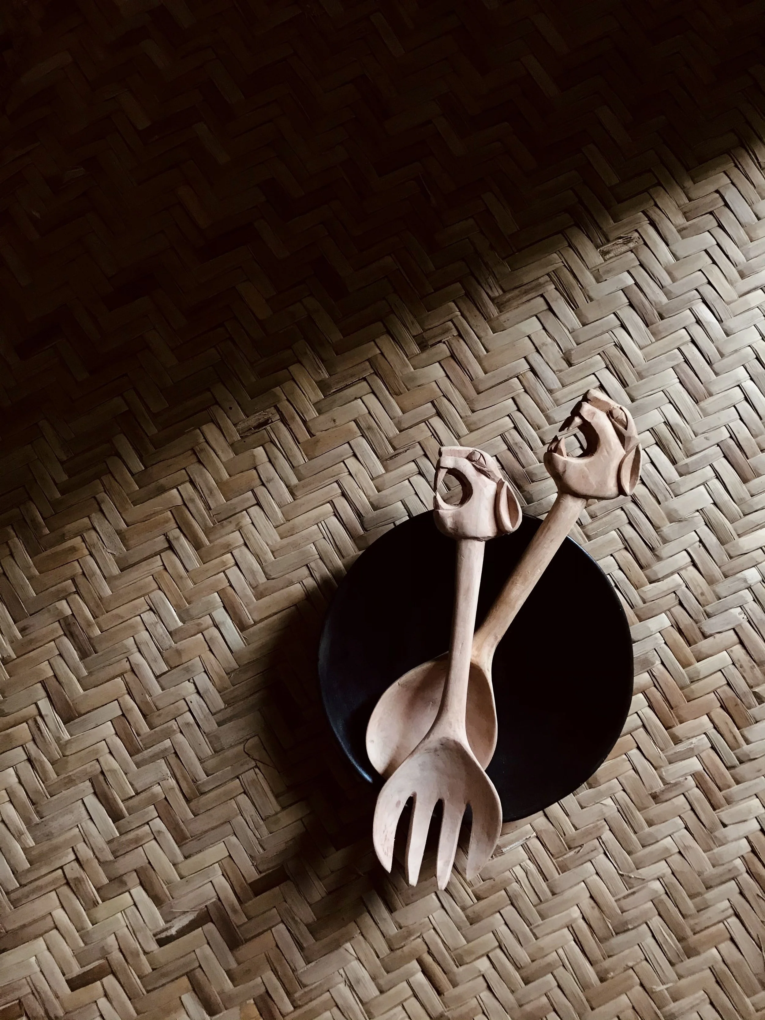 Deux cuillères en bois avec des têtes sculptées en forme de tête de chien, placées dans un bol noir sur un tapis en osier.