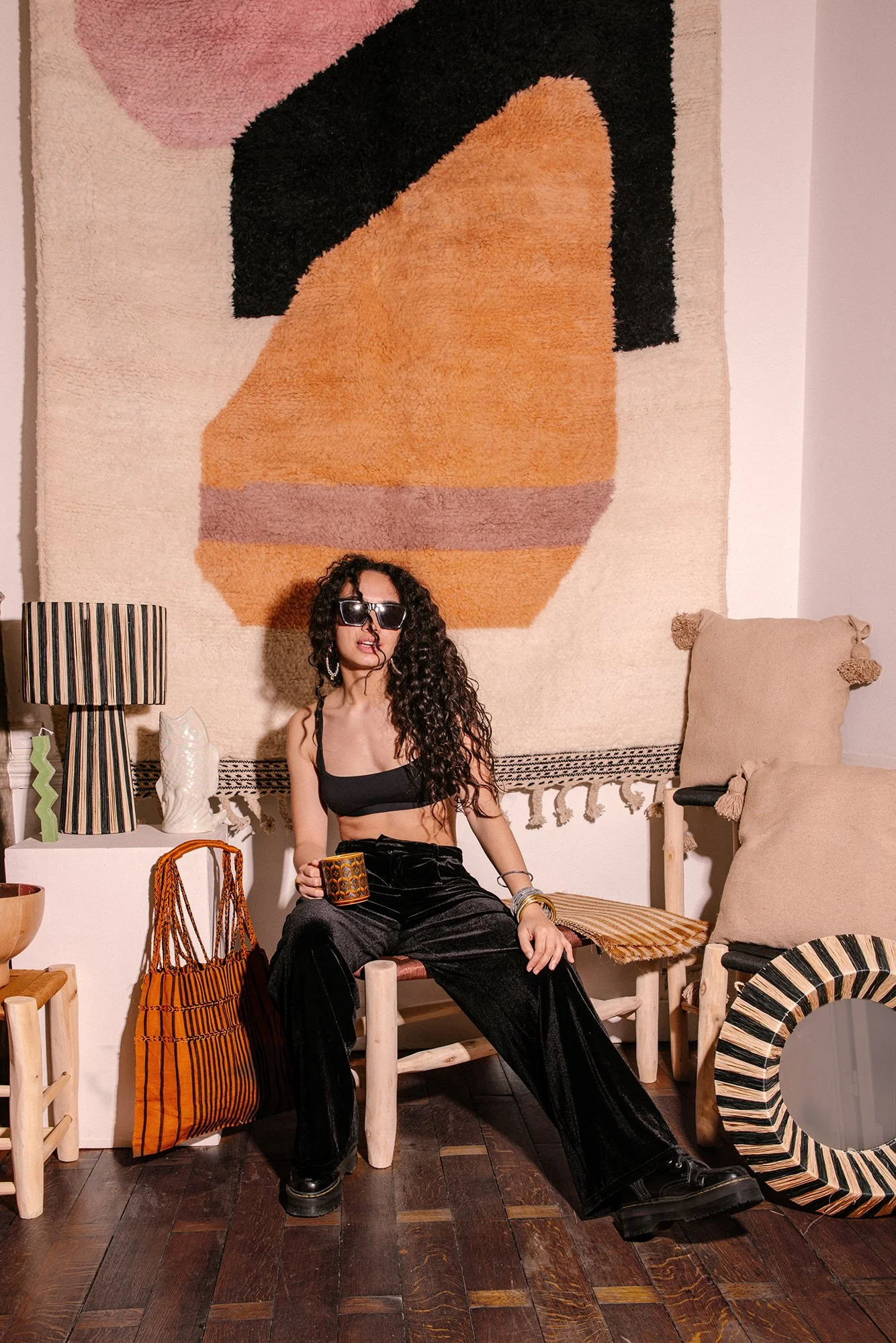 Une femme assise dans une pièce décorée de façon moderne, portant des lunettes de soleil noires, une robe de blouse noire, des pantalons en velours noirs, et tenant une tasse. En arrière-plan, un tapis mural abstrait, un lampadaire à rayures, des coussins beige, et un miroir rond.