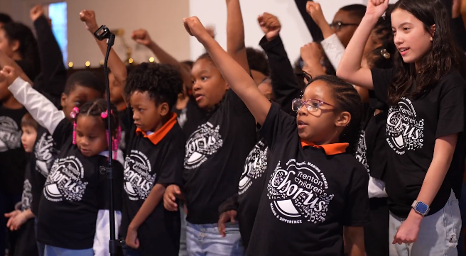 Children wearing matching black t-shirts participating in a group activity, raising their fists, in a room with a microphone stand.