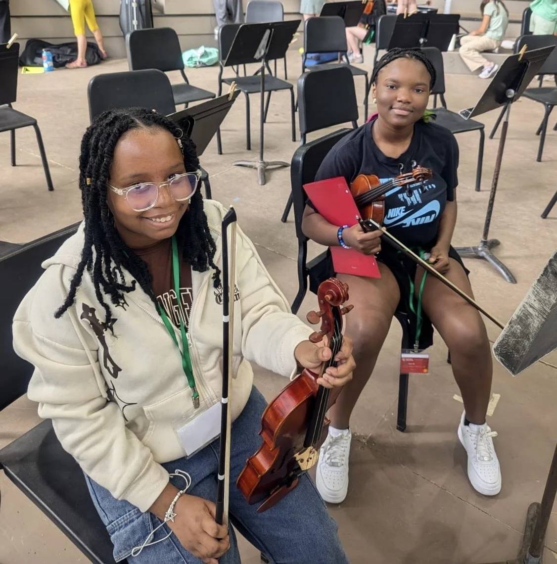 Two young girls holding violins and bows, sitting in a room with music stands and chairs, likely at a music class or rehearsal. The girl on the left has glasses and dreadlocks, and the girl on the right is wearing a Nike t-shirt, holding a sheet of music.