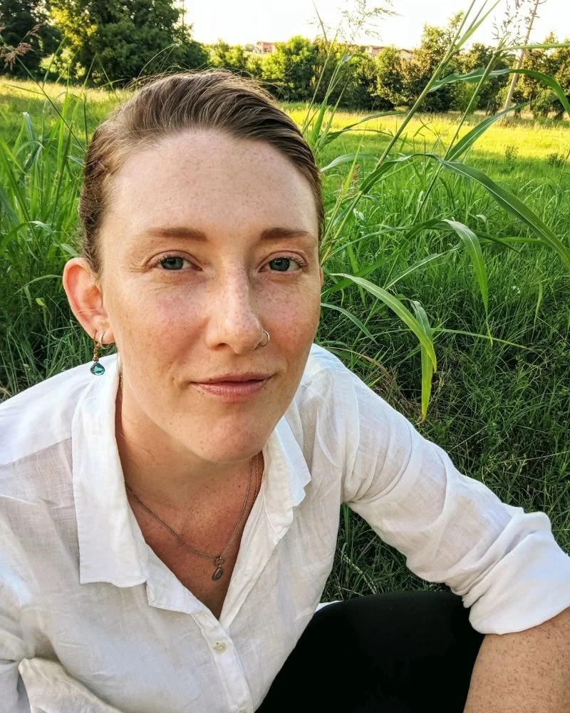 Muse, Jen Aliz Lokajickova looks at the camera. She is wearing a white button down linen shirt and blue topaz earrings. She sits in the grass outside a village in Italy.