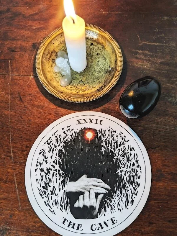 the cave card from the archtypes deck by kim krans, on a wooden table, next to an onyx egg and a white candle with wax pooling at it's base, casting a glow across the photo