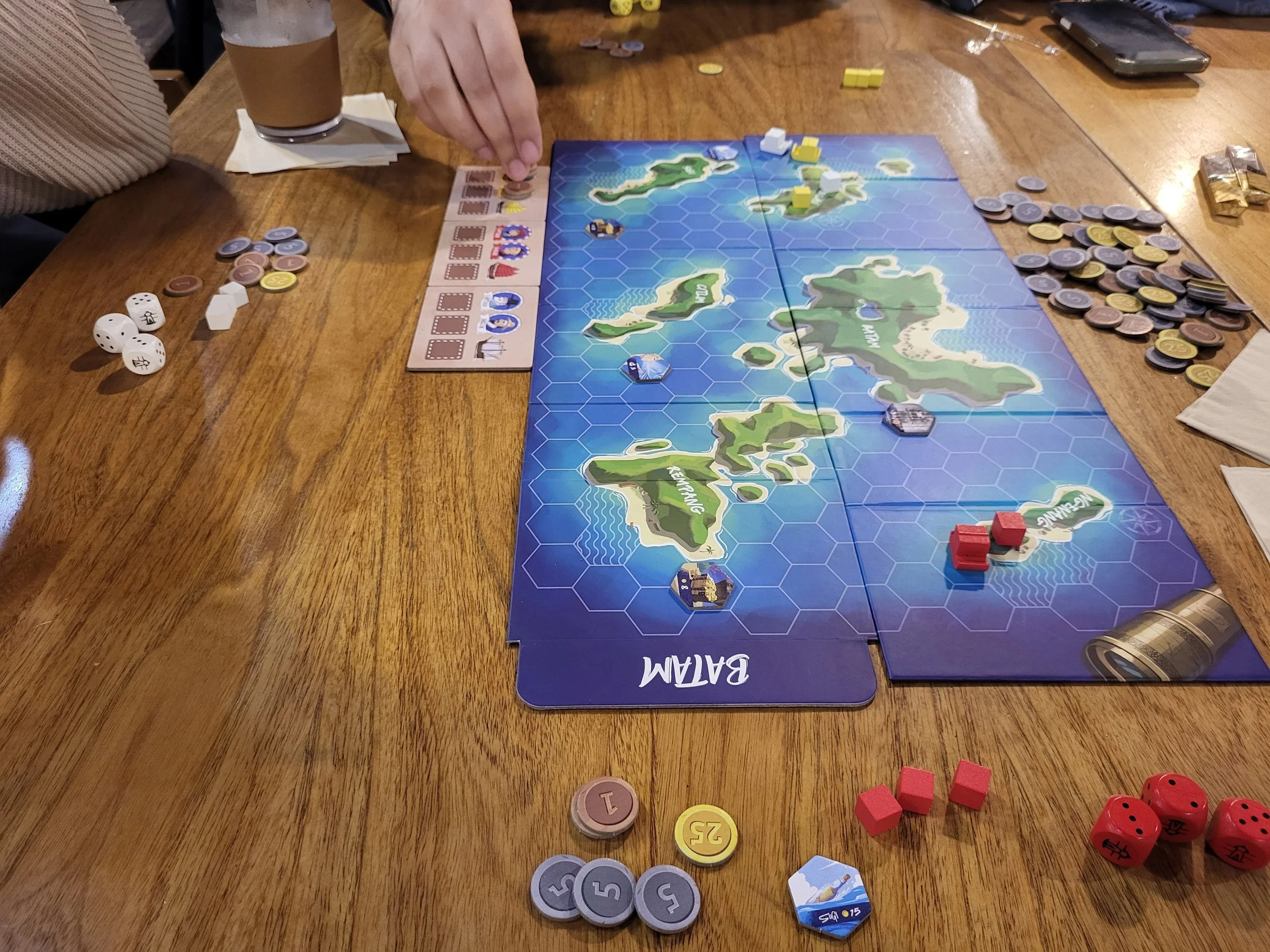 A photo looking down at the board of Batam from the eastern edge. A hand is reaching into frame to place coins.