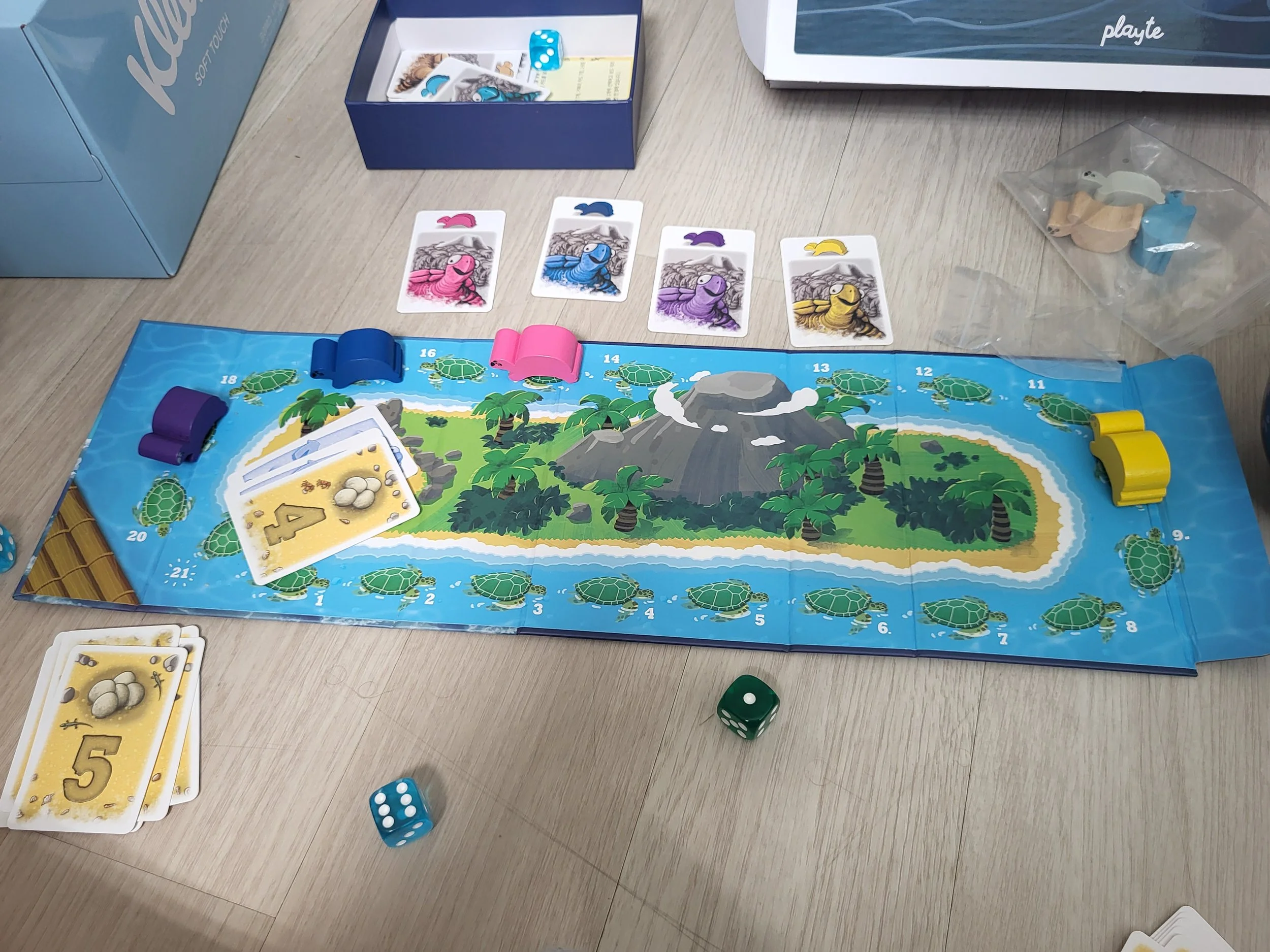 The middle of a game of Mahe, with four colored wooden turtles on the folded out board. Cards show the order the turtles move in, and there is a pile of scoring cards.