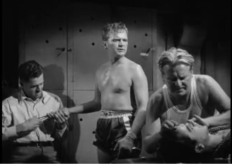 Four people in a locker room. On the left a guy is looking after the hands of the boxer in the center, while in the front we see the head of one recovering boxer (lying on a table out of frame) being taken care of by another man.