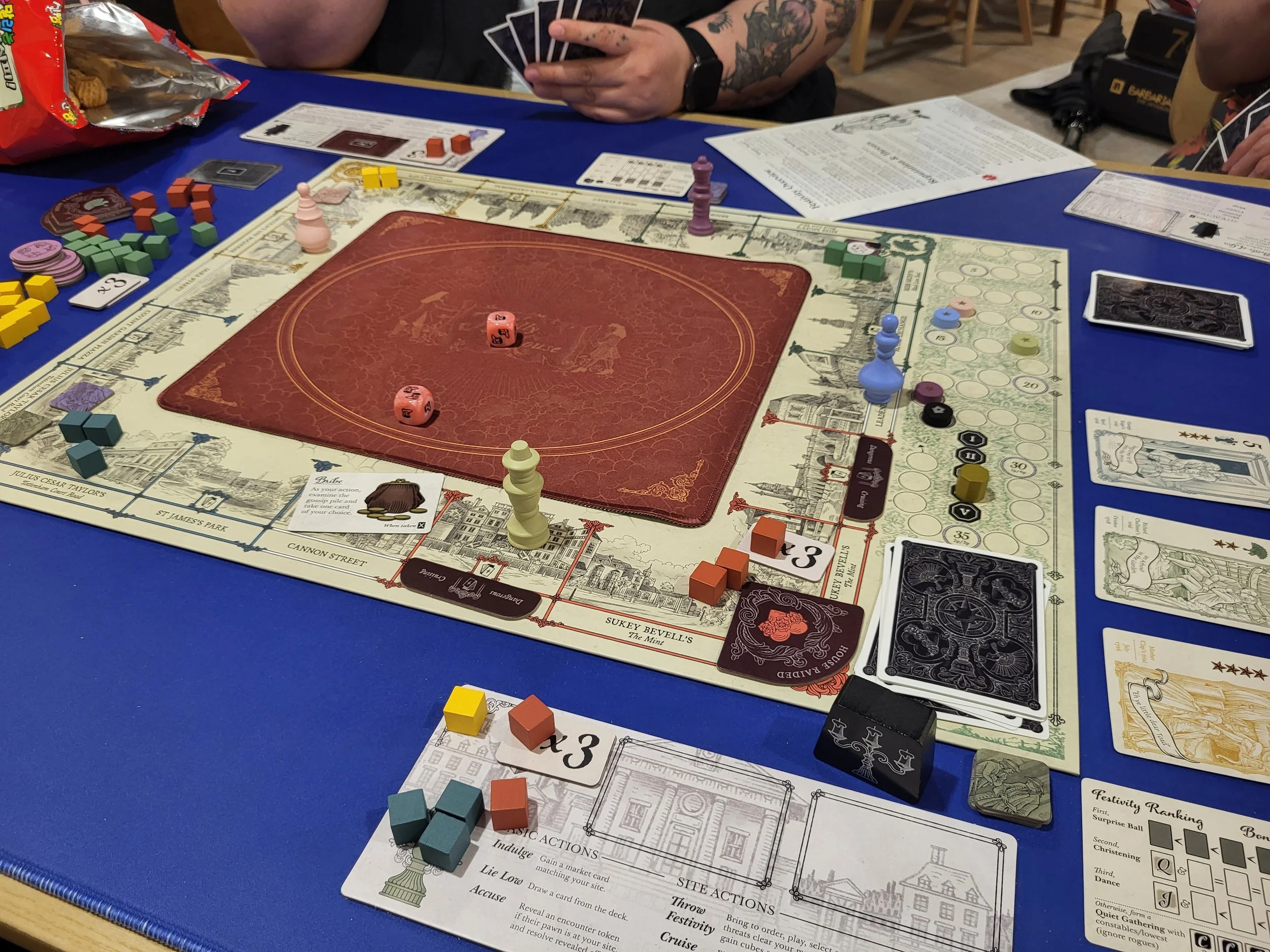 A view of a game of Molly House in progress. My player board is close to the camera, another player's arm appears in the top, but the main focus is the board and cards.