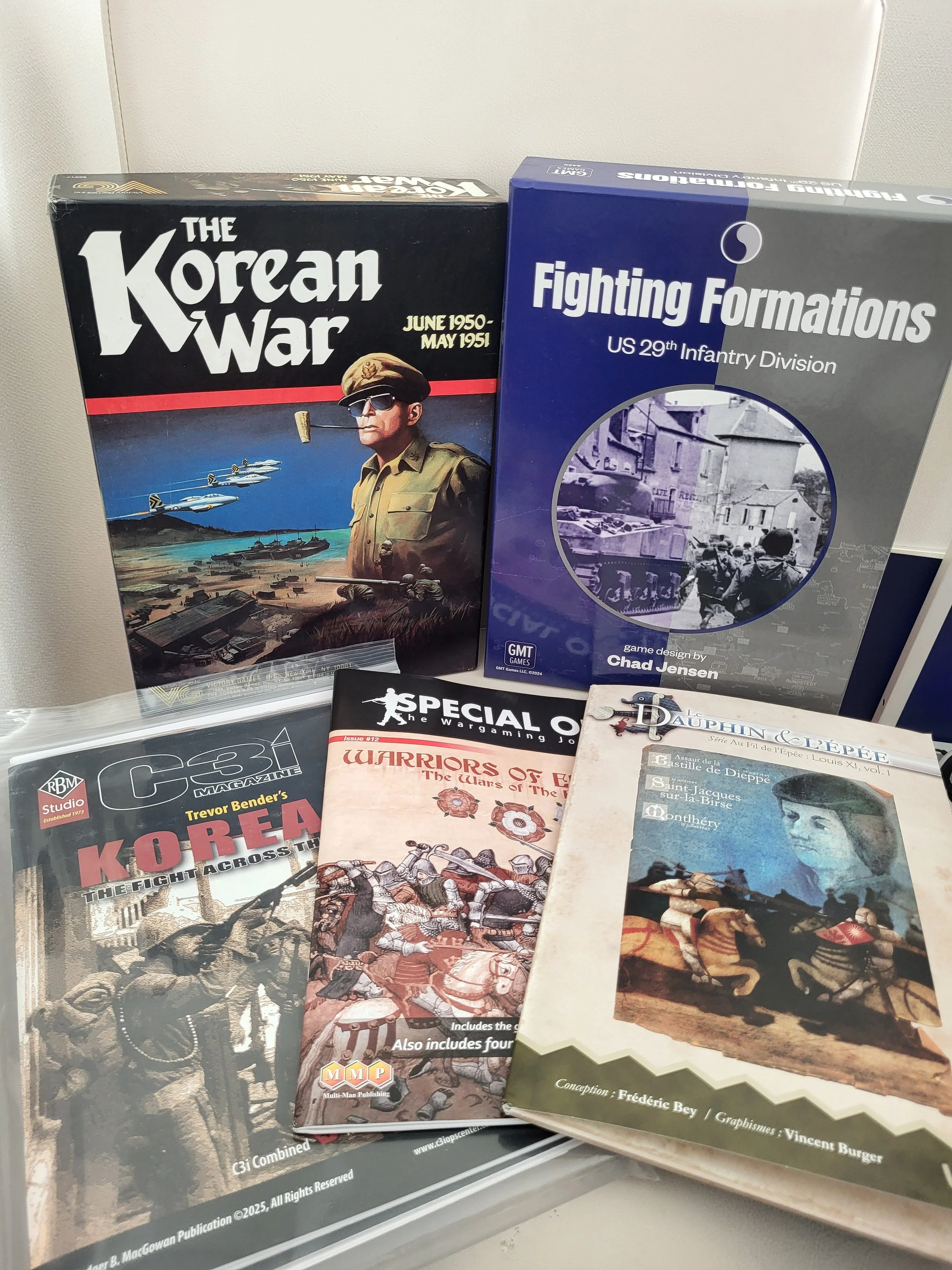 The Victory Games edition of Joseph Balkoski's The Korean War next to Chad Jensen's Fighting Formations with three folio/magazine games (Trevor Bender's Korea in C3i, Warriors of England in Special Ops, and Dauphin & l'Epee) all laid out on a chair