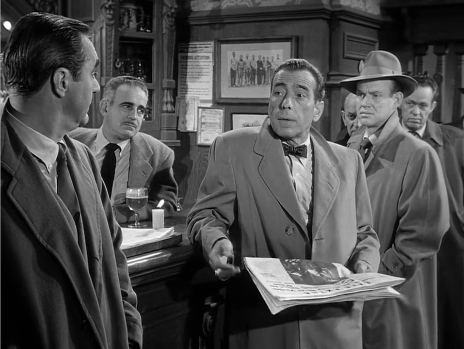 It's a group of serious looking dudes in a bar, Bogart is leaning on the bar with his coat on and holding a newspaper. He's clearly mid-speech trying to persuade the grumps around him