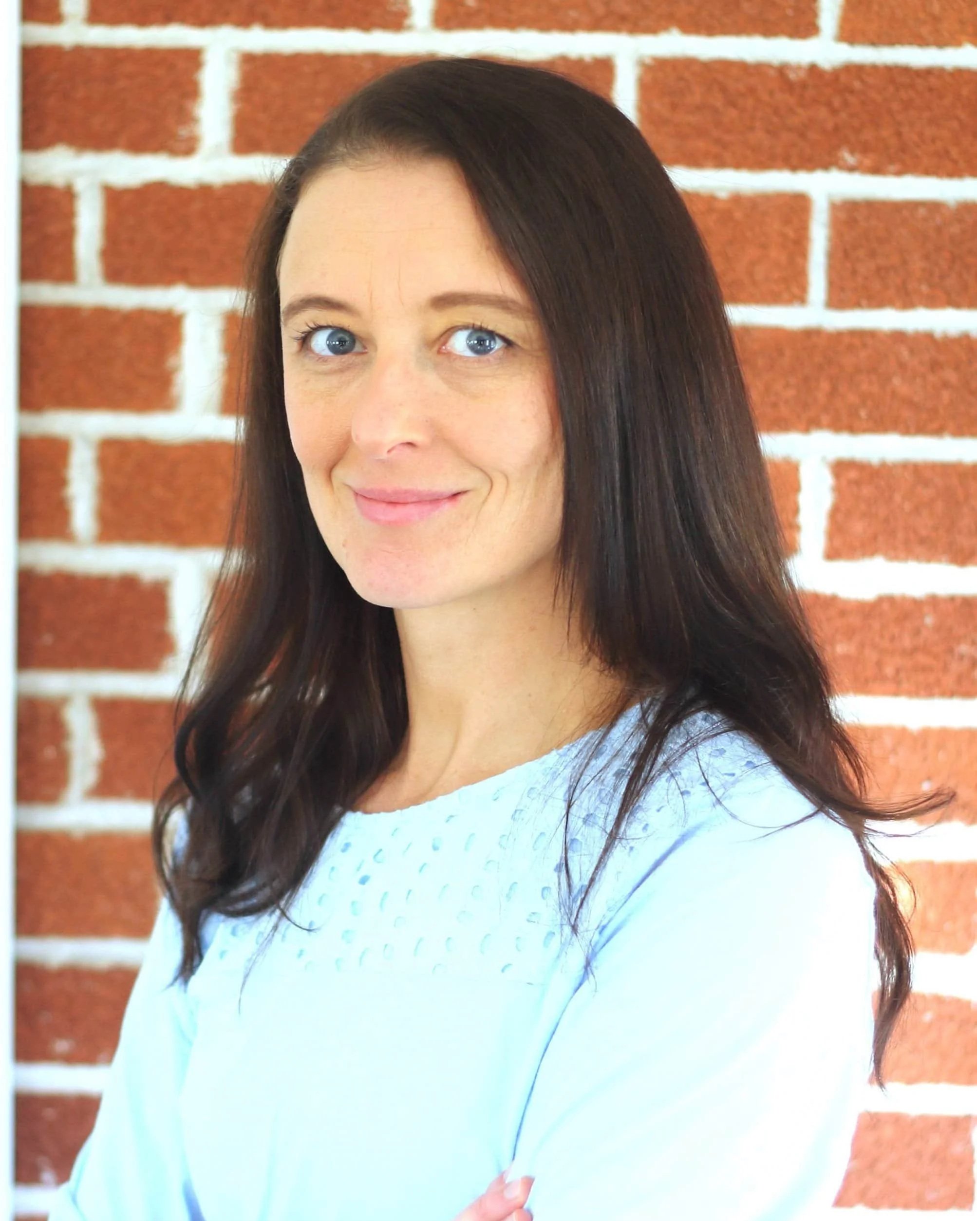 A woman with long dark brown hair and blue eyes, wearing a light blue top, standing in front of a red brick wall, smiling at the camera.