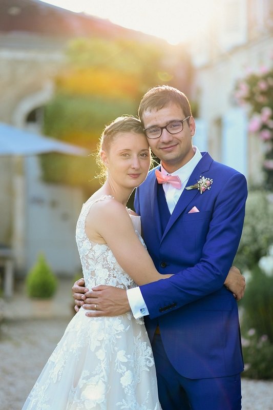 Marie et Fabien sont face à face enlacés. Le marié embrasse sa femme sur la joue. La lumière du soleil couchant en arrière plan met en valeur la robe et le costume.