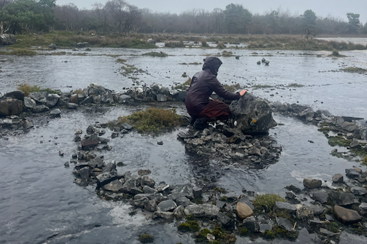 A person sitting on a rock in the middle of a shallow river, fishing.