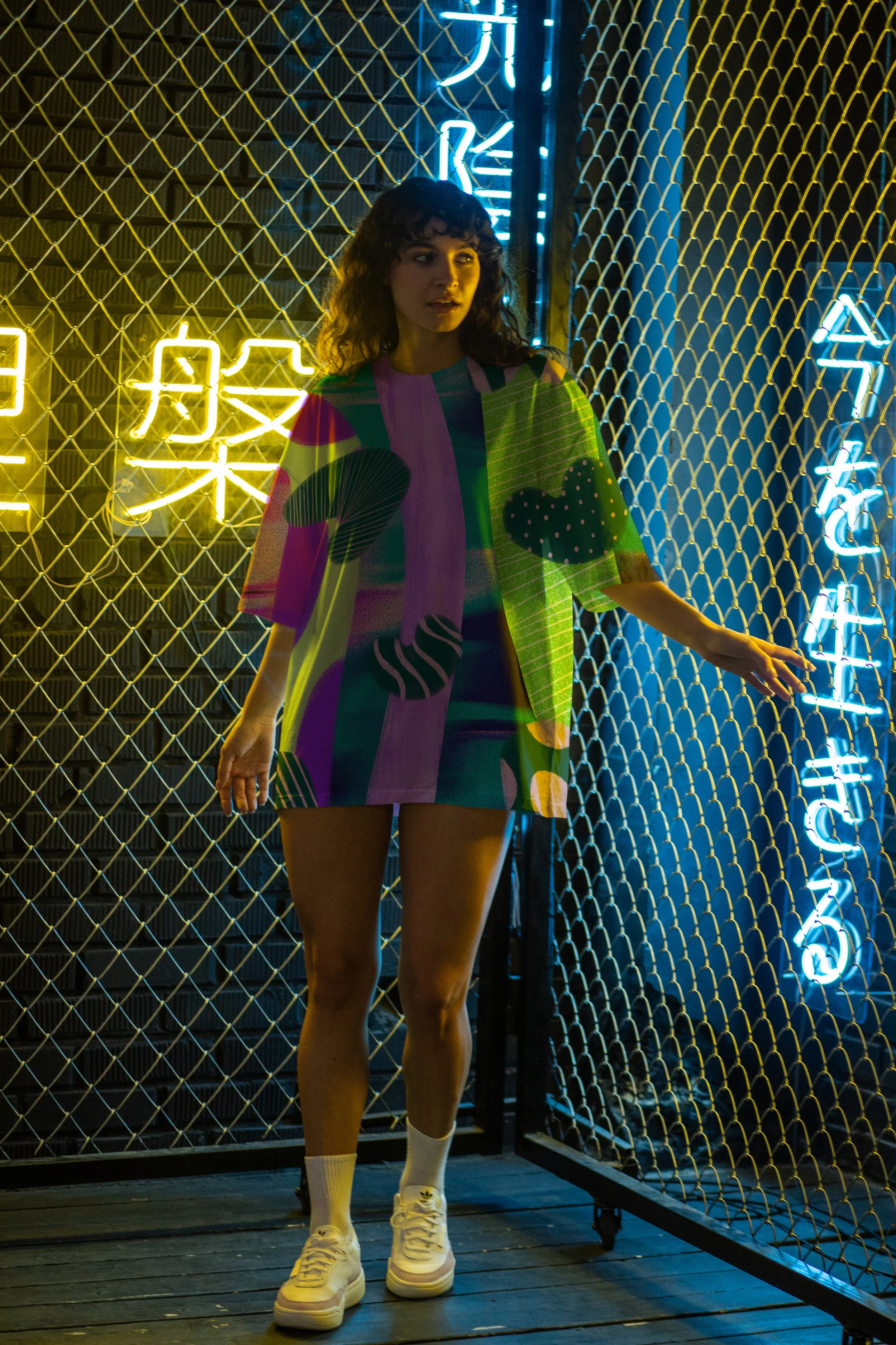 A woman standing in front of a chain-link fence with illuminated neon signs in the background, wearing a colorful oversized shirt and white sneakers.