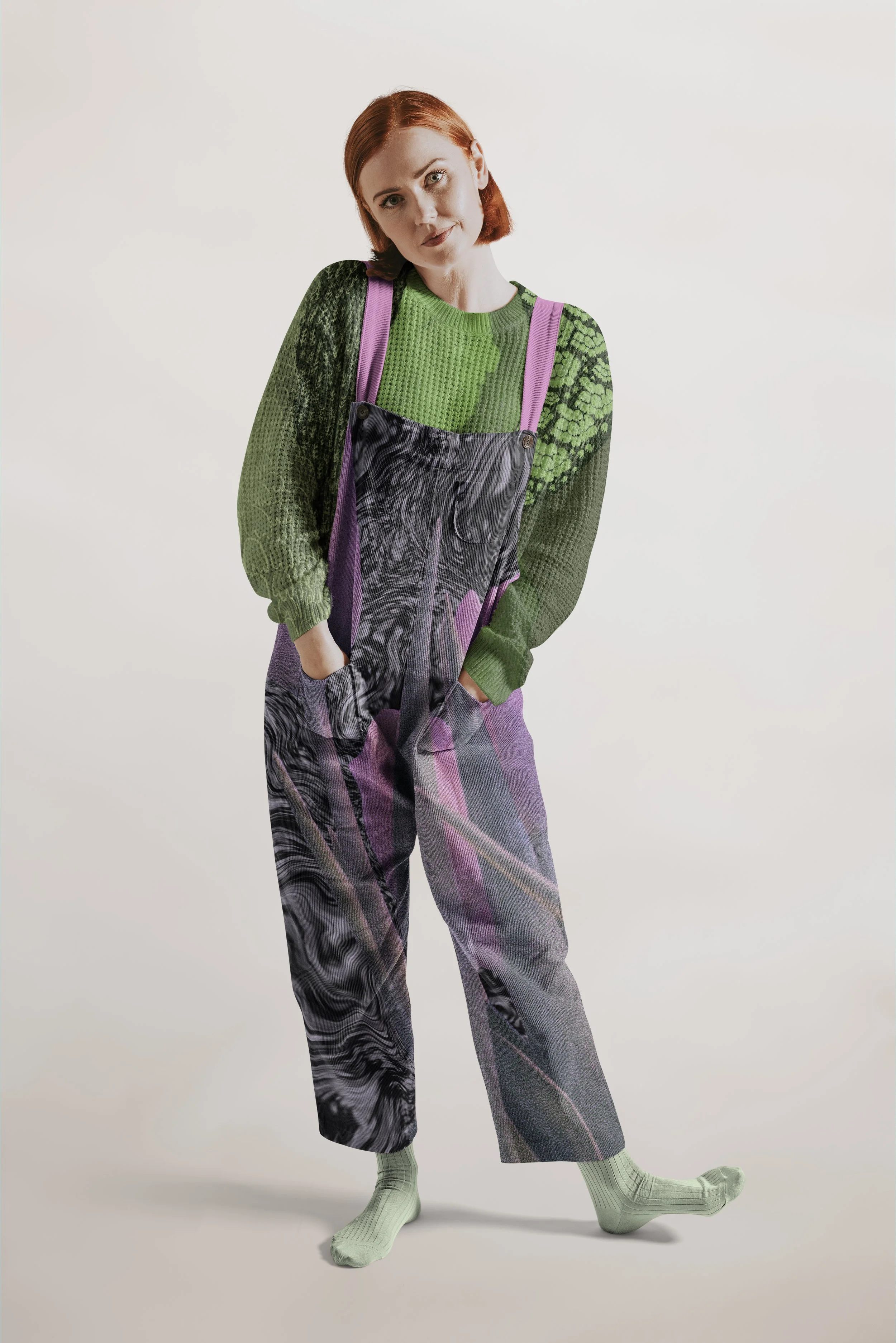 A person with red hair wearing a green sweater with semi-sheer pattern, black and purple overalls with a swirling design, and pale green socks, standing against a plain light background.