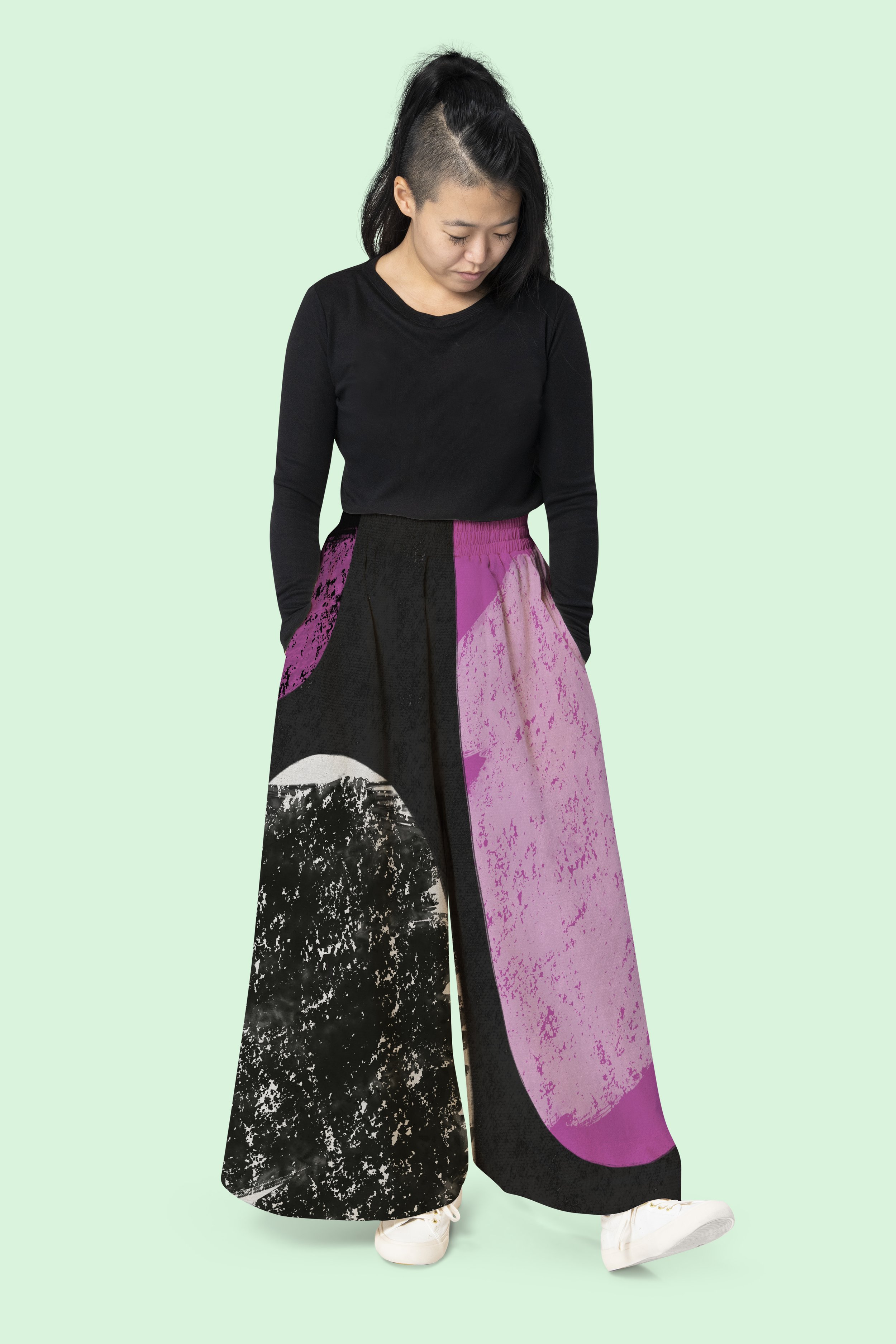 A woman wearing a black long-sleeve shirt and wide-leg pants with a pink, black, and white abstract pattern, standing against a light mint green background.
