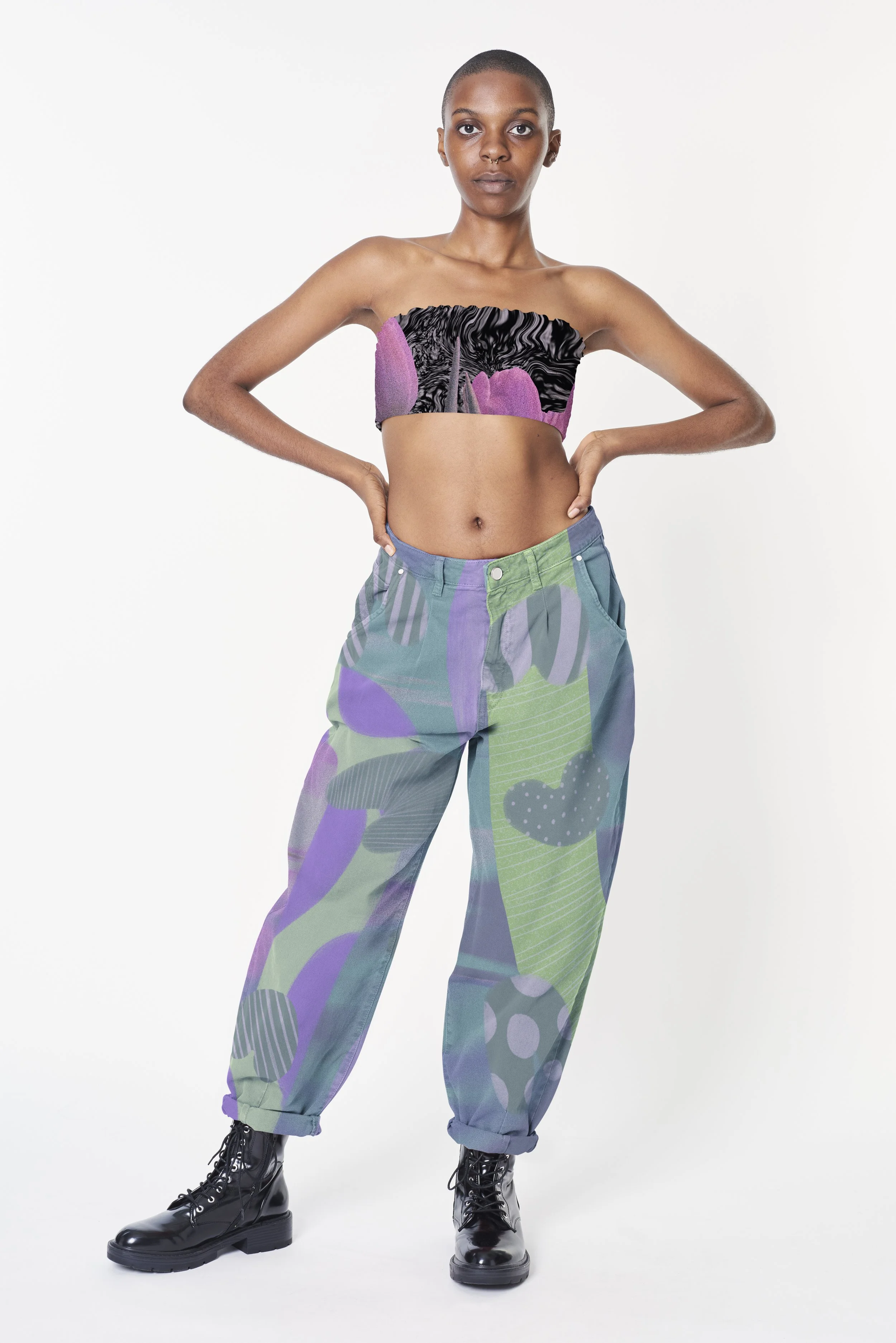Person with short hair wearing a black and purple patterned tube top, loose multicolored patchwork jeans, and black platform boots against a plain white background.