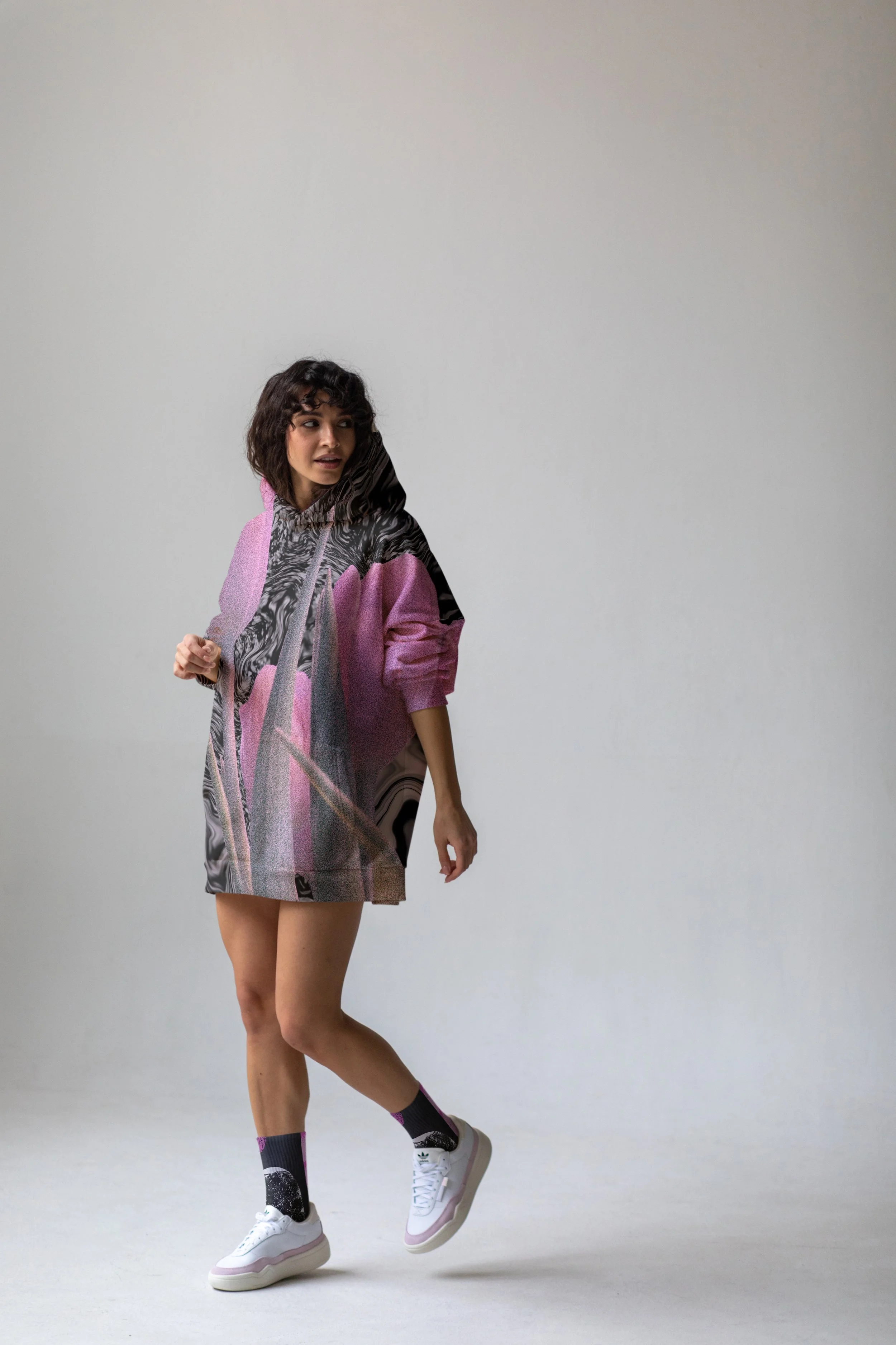 A young woman with curly dark hair wearing a colorful, patterned hoodie, shorts, and white sneakers, standing against a plain gray background.
