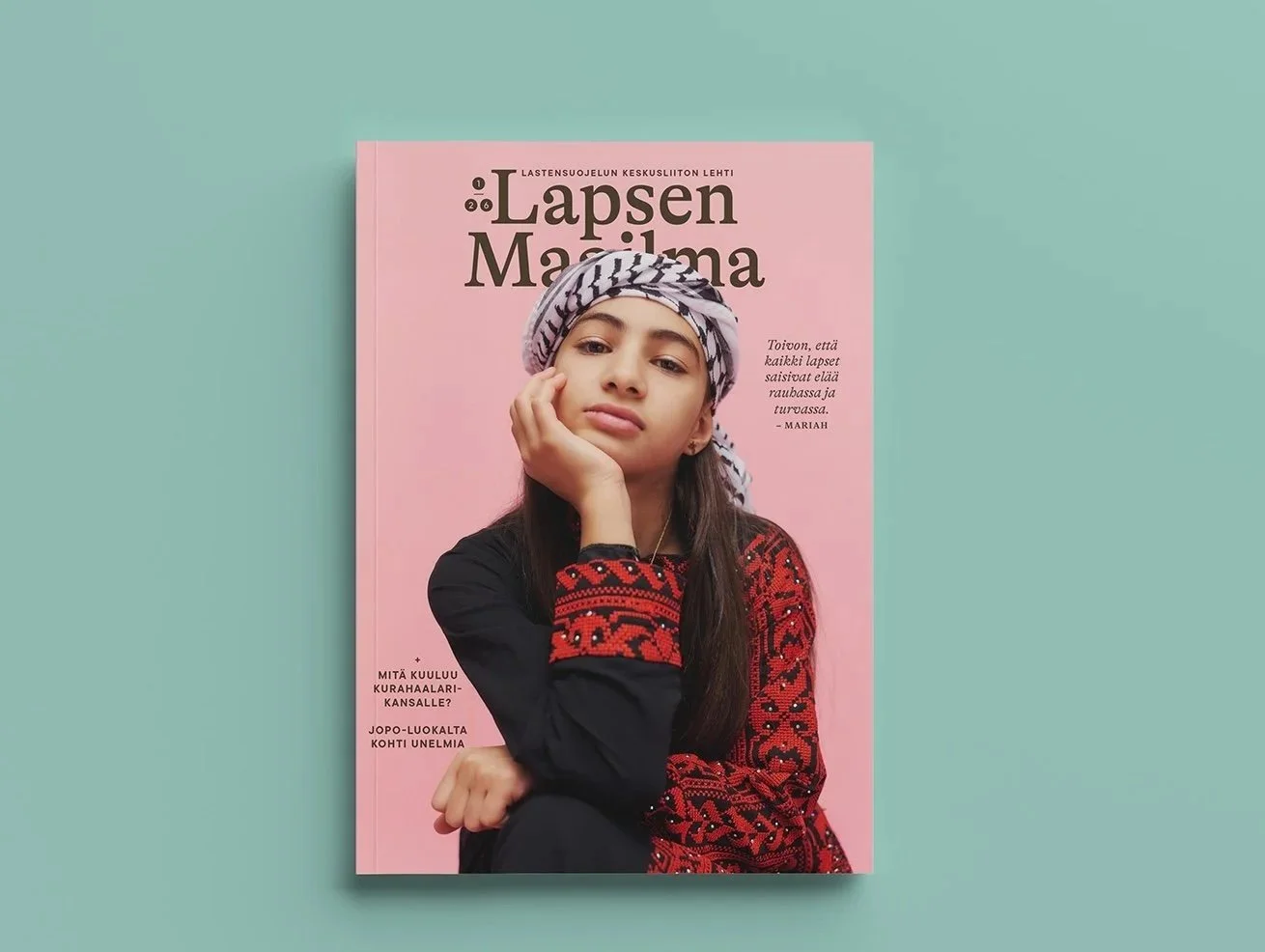 Nuoreen nuotti, pää pään ympäri kääritty sinisellä ja valkoisella pyyhkeellä, musta ja punainen vaatetus istuu pinkillä taustalla olevaan lehtikuvakkeeseen, jossa lukee 'Lapsen Maailma'.